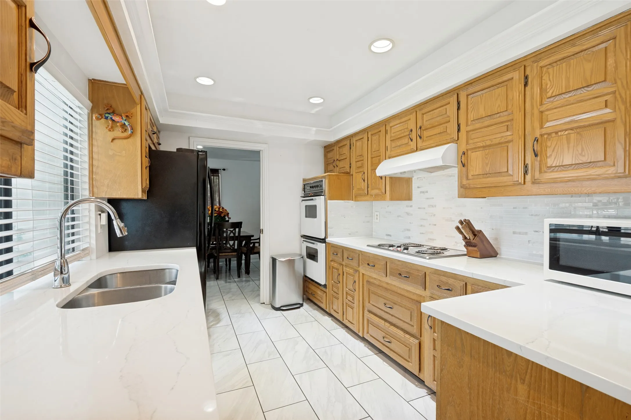 Galley kitchen featuring light wood-finish cabinetry, white countertops, stainless steel double basin sink, white tile backsplash, and recessed lighting