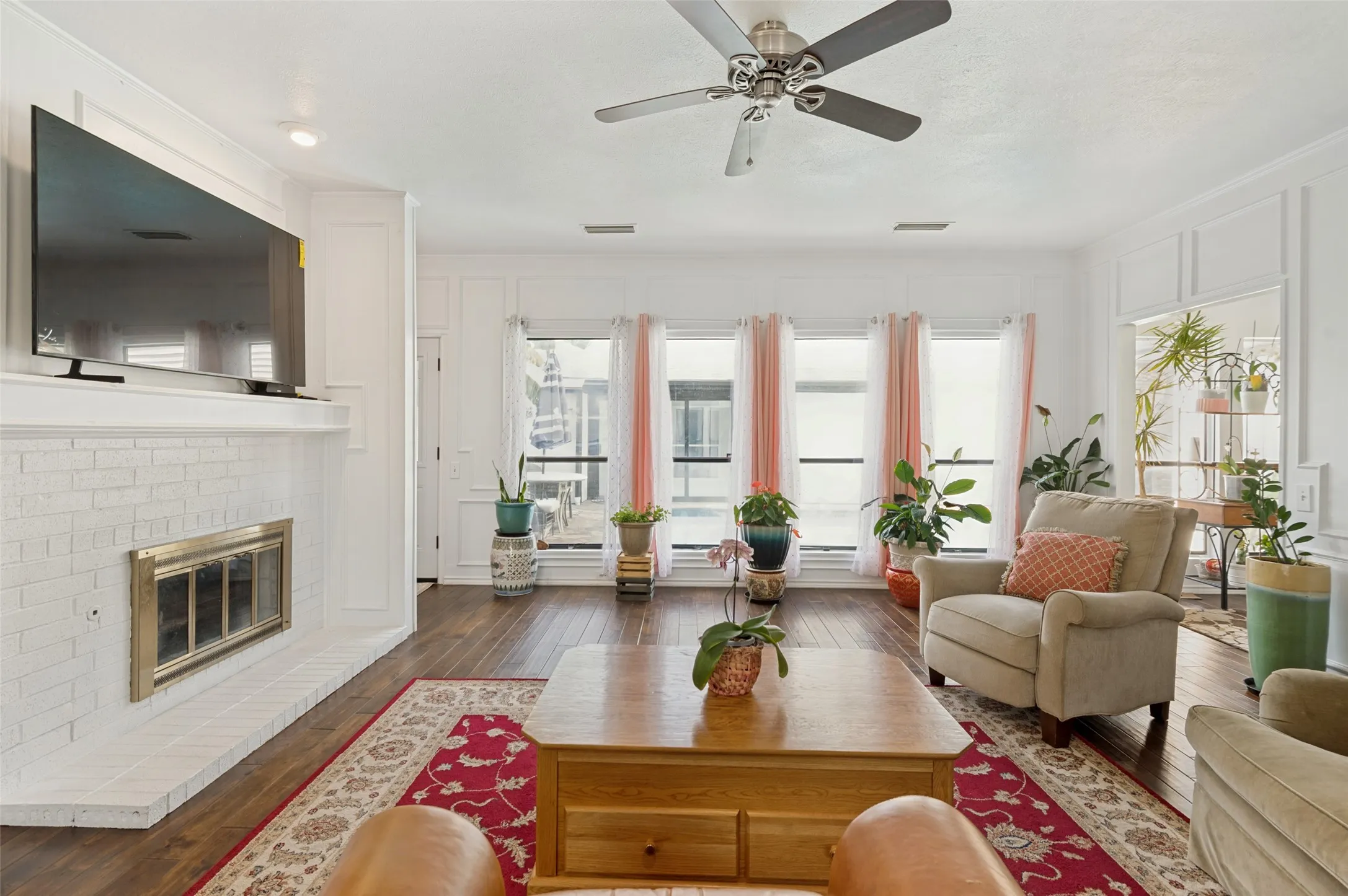 Living area featuring wood-finish flooring, a white painted brick fireplace with a metal insert, a ceiling fan with integrated lighting, recessed lighting, and multiple windows