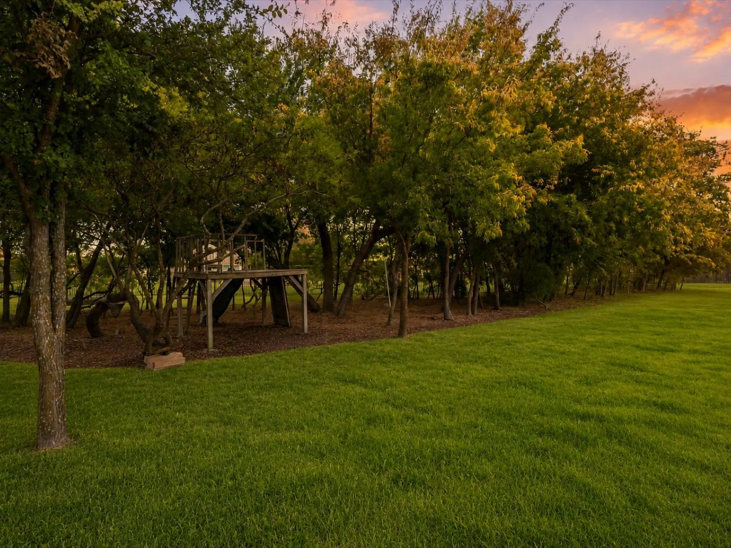 Expansive green lawn with a tree-lined perimeter, featuring a multi-level wooden play structure