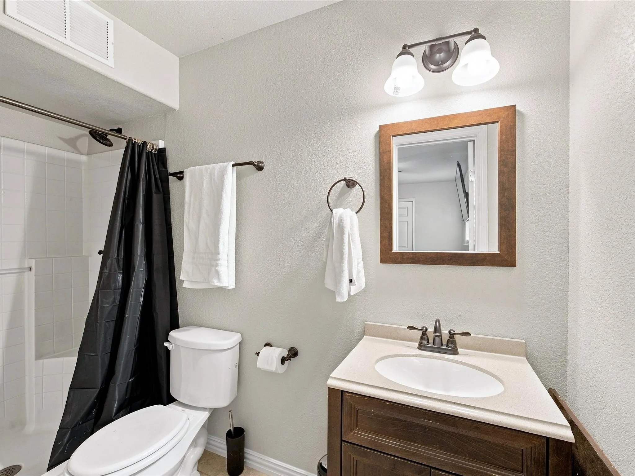 Shower with white subway tile, vanity with integrated sink and wood-finish cabinetry, framed mirror, and two-light vanity fixture