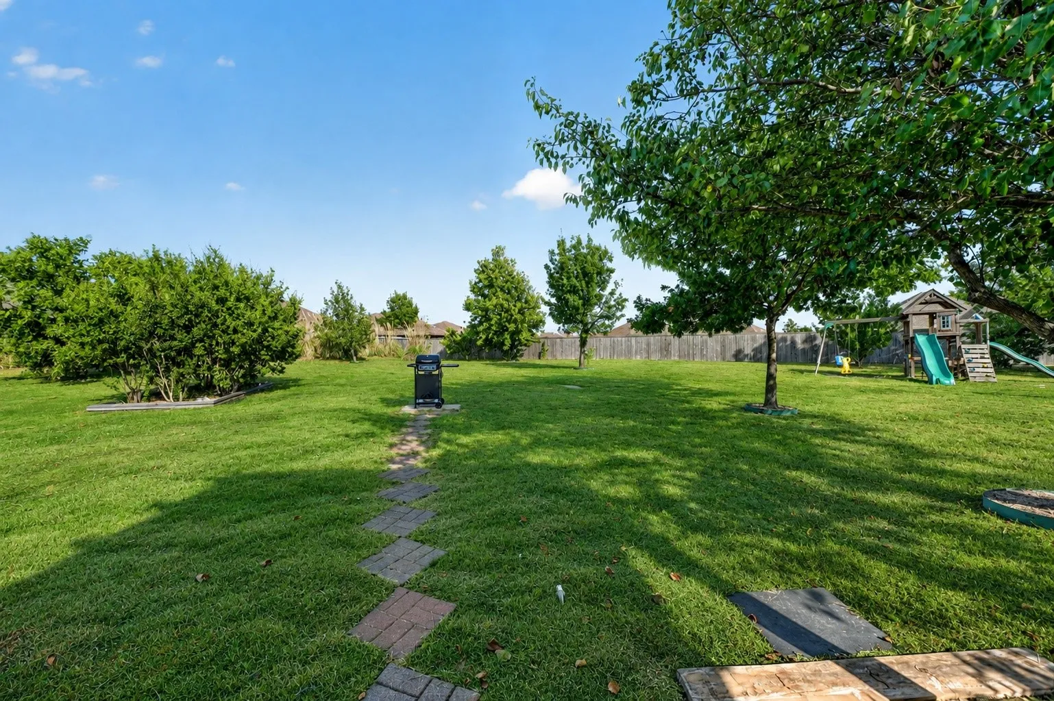 Expansive backyard with mature shade trees, manicured lawn, paver pathway, children's play structure, and storage shed