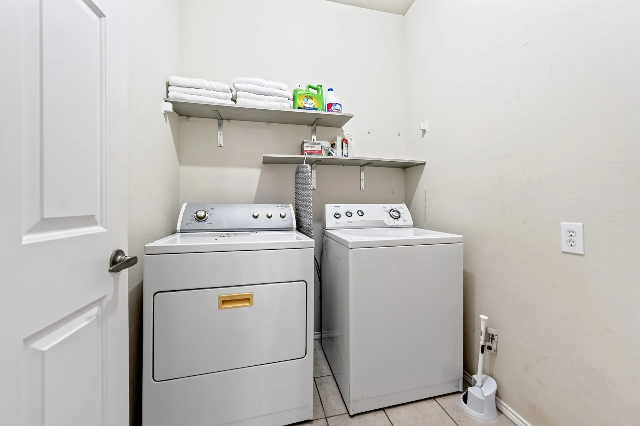 Dedicated laundry room featuring side-by-side front-load washer and dryer with tile flooring, wall-mounted shelving for storage, and convenient utility sink