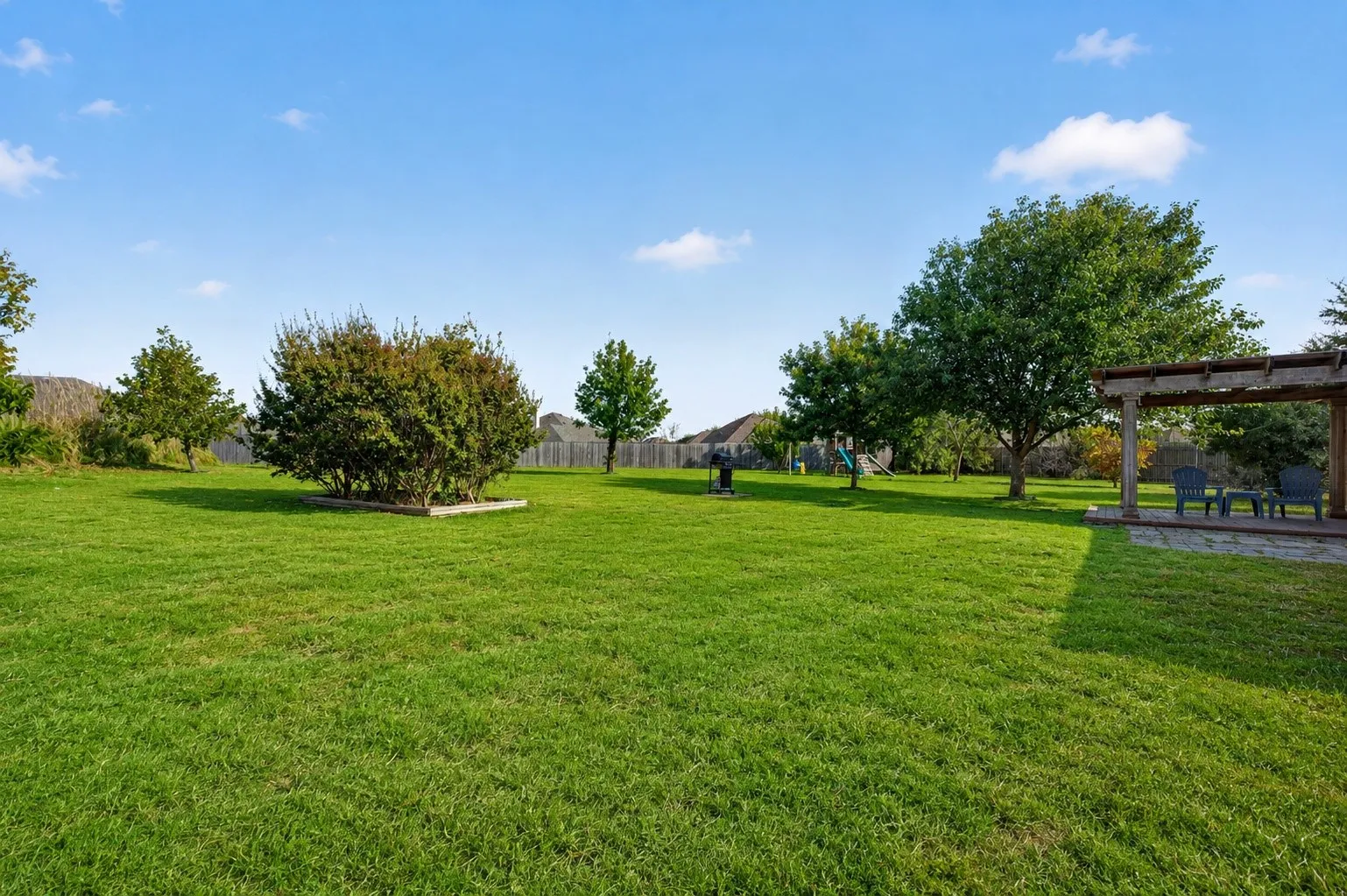 Expansive manicured lawn with mature shade trees and open grounds, complemented by a covered outdoor structure and hardscaping features visible in the distance