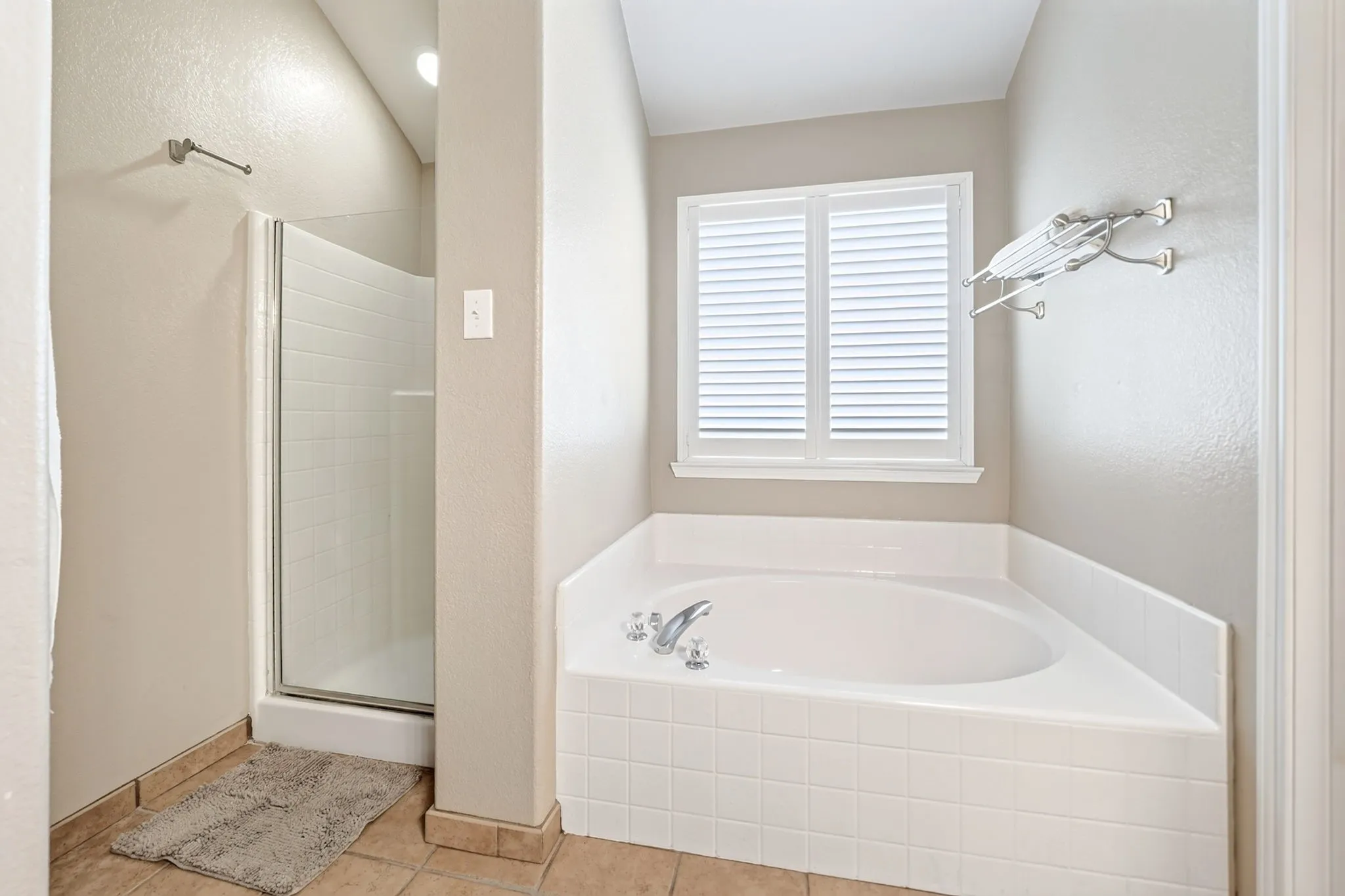 Spa-inspired primary bathroom featuring a curved soaking tub with dual faucet fixtures, a walk-in shower with frameless glass enclosure and rainfall showerhead, ceramic tile surround, plantation shutters, and tile flooring throughout