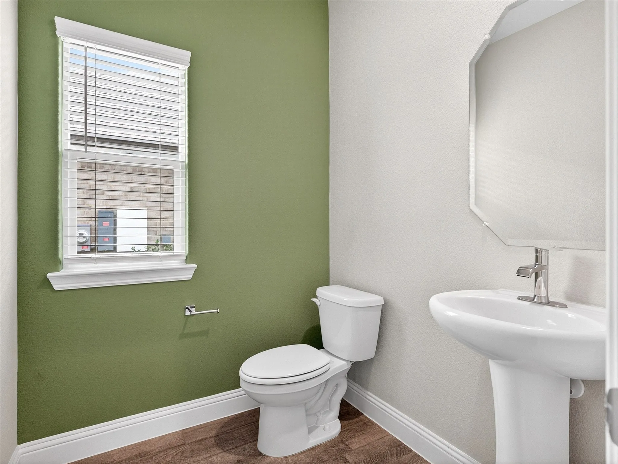 Pedestal sink with chrome-finish faucet, elongated toilet, wood-finish flooring, and a window with blinds