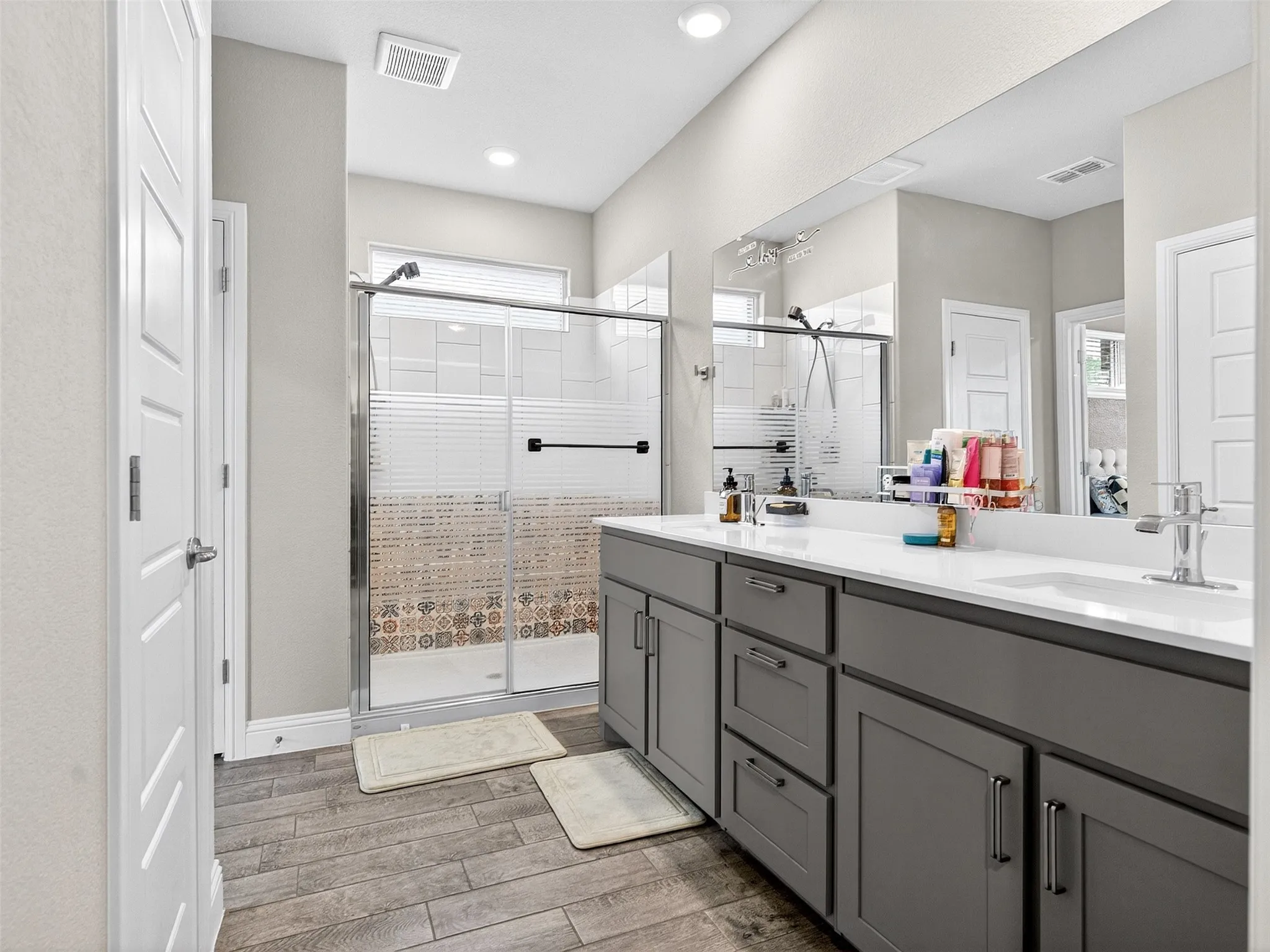 Dual vanity with under-sink storage, wood-finish flooring, and a glass-enclosed shower with decorative tiling