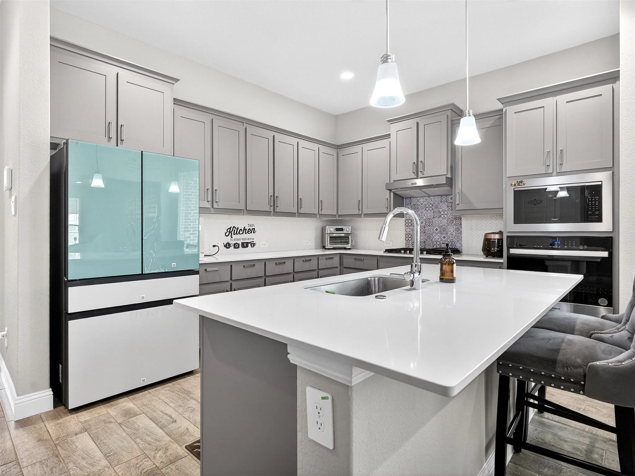 Kitchen featuring gray cabinetry, white countertops, stainless steel appliances, a center island with an undermount sink, and wood-finish flooring