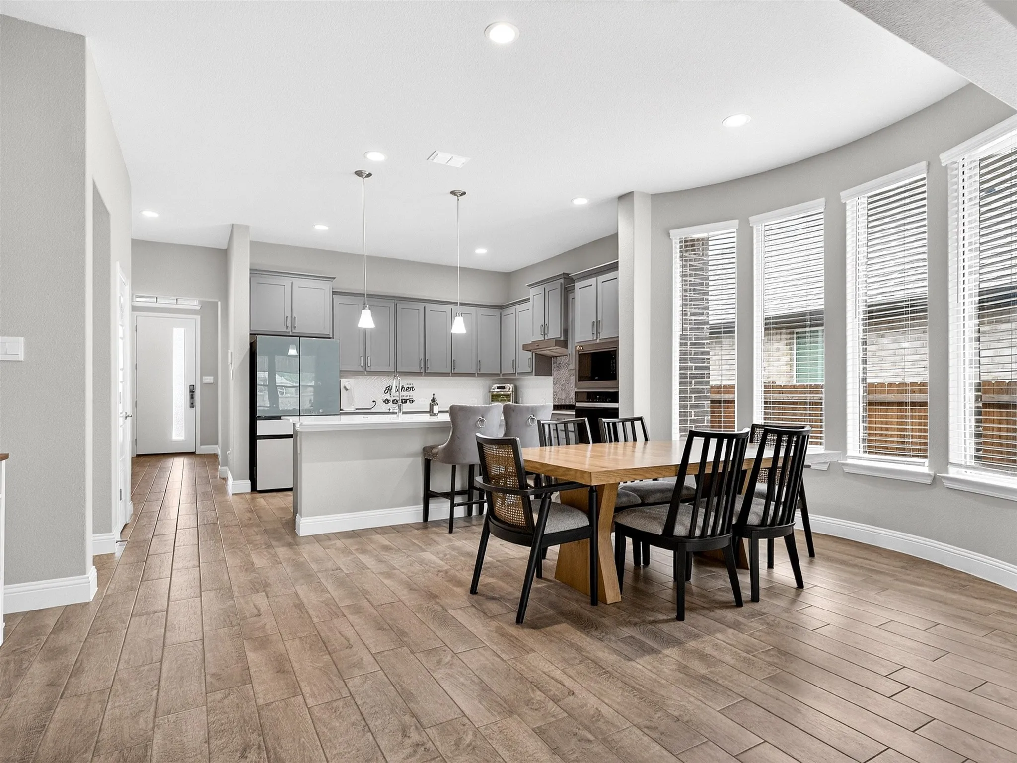Wood-finish flooring throughout, curved wall with multiple windows, recessed lighting, and kitchen featuring gray cabinetry and pendant lights