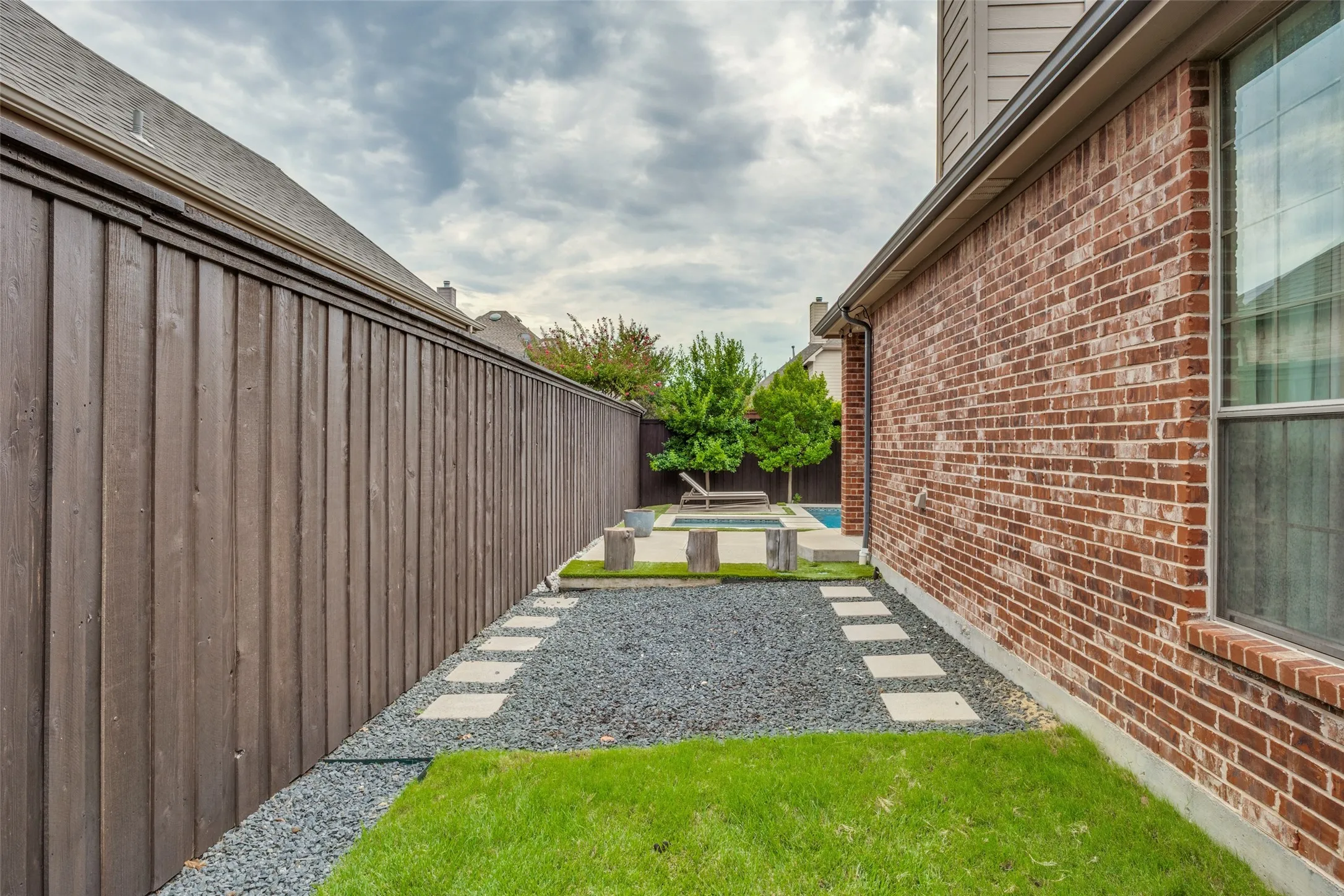 Brick exterior with a large window, privacy fence, and a landscaped side yard featuring a grass lawn, gravel path, and stepping stones