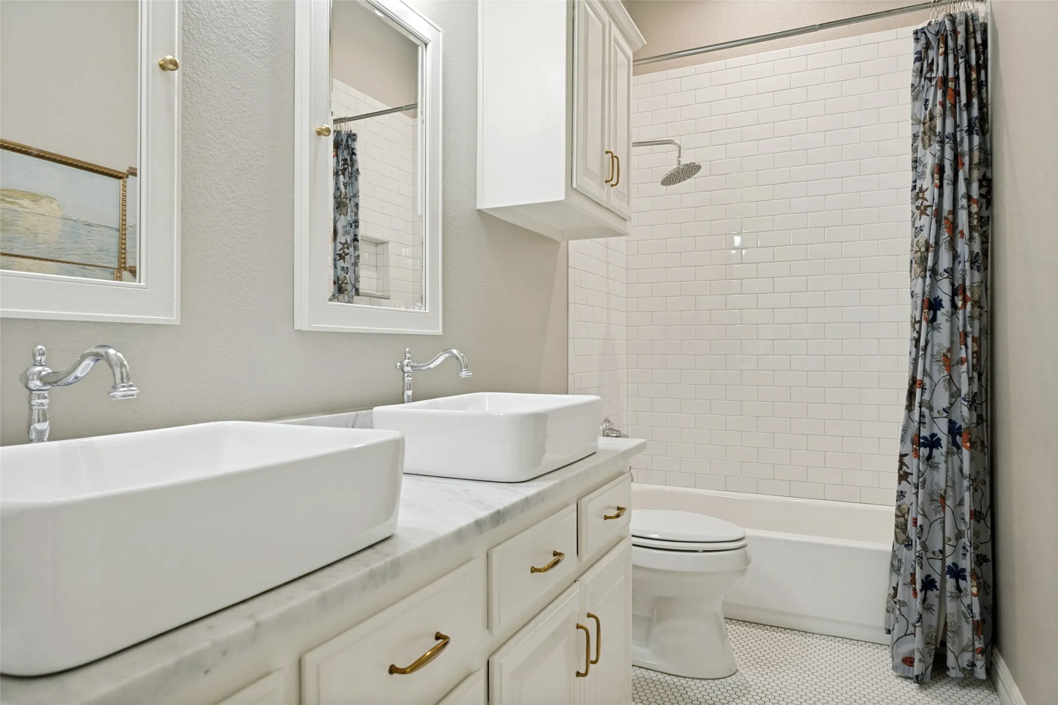 Dual vessel sinks set on a marble-finish countertop, white cabinetry with brass-finish hardware, subway tile shower surround, and hexagonal tile flooring