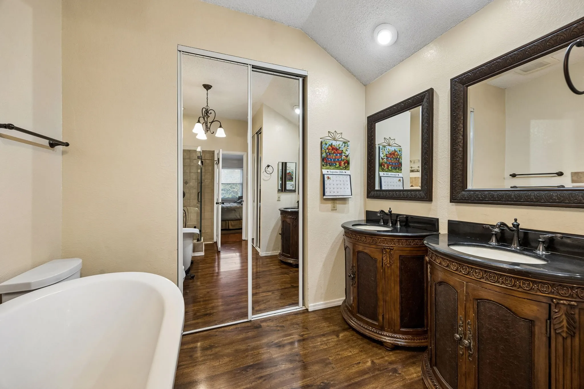 Freestanding white soaking tub, wood-finish flooring, dual vanity with dark countertops, wood-finish cabinetry, and mirrored sliding closet doors