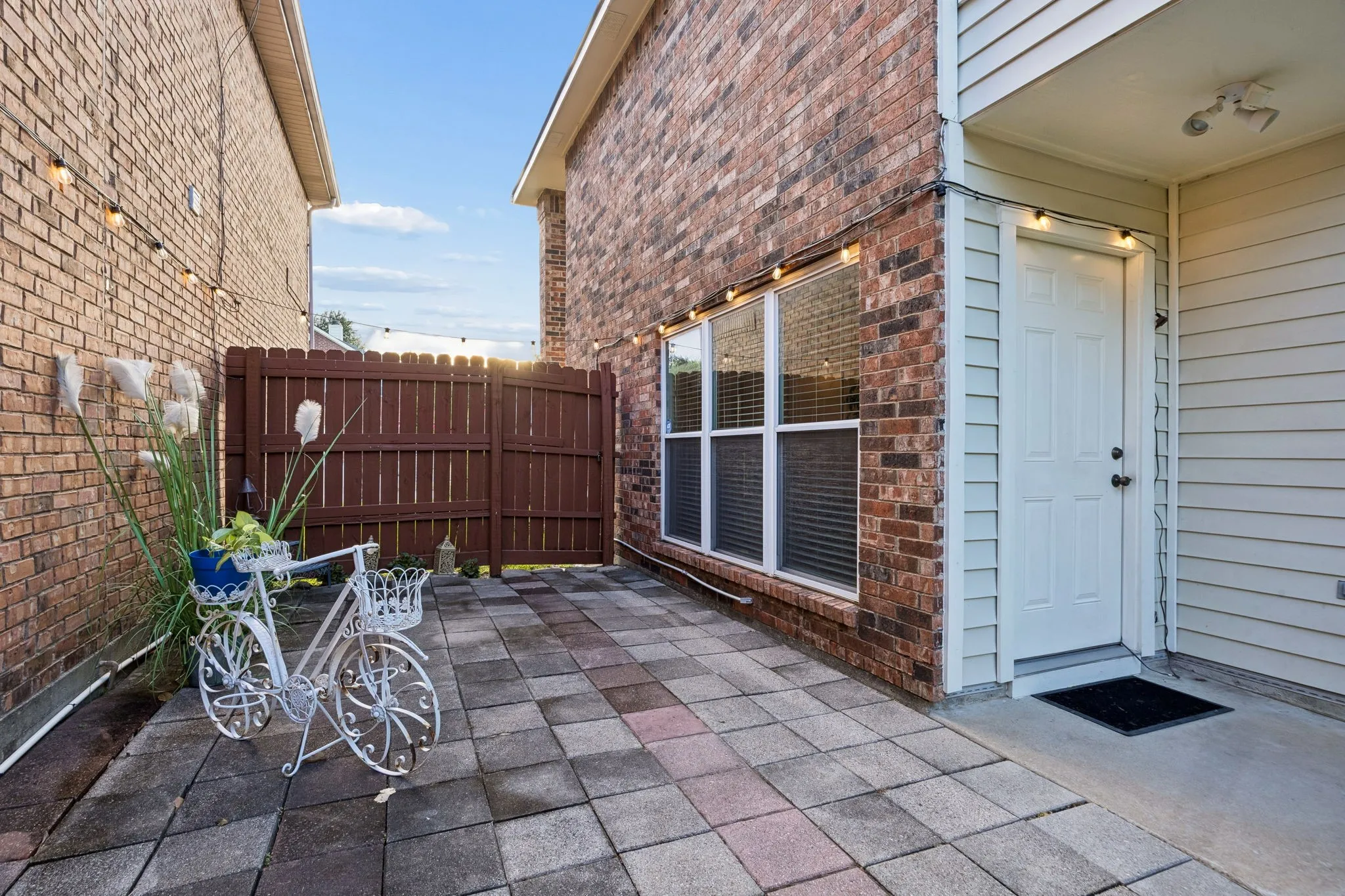 Paved outdoor patio with brick and siding exterior, privacy fencing, and exterior lighting fixtures