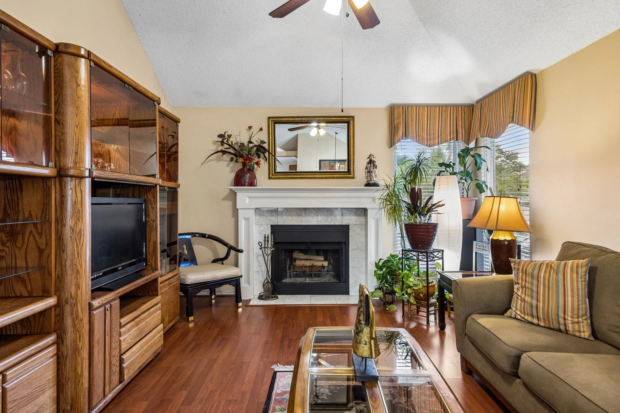 Vaulted ceiling with ceiling fan, wood-finish flooring, fireplace with white mantel and stone surround, and large windows with blinds