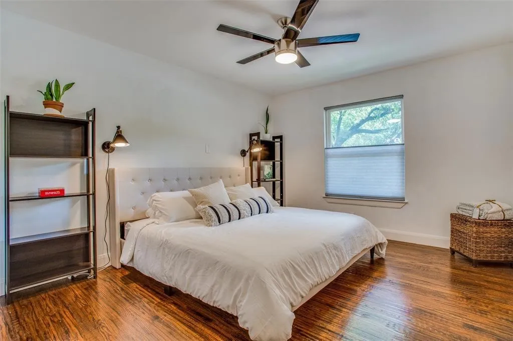 Hardwood flooring, ceiling fan, window with privacy shades, built-in wall sconces, and a tufted headboard