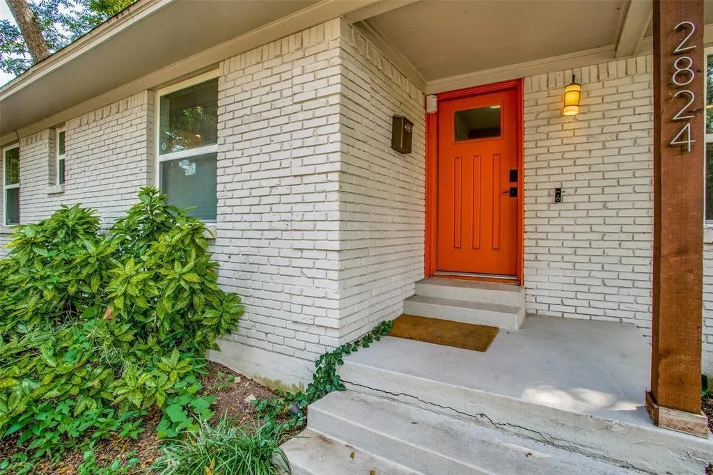 White brick exterior with an orange entry door, concrete steps, and a wood-finish column with house numbers