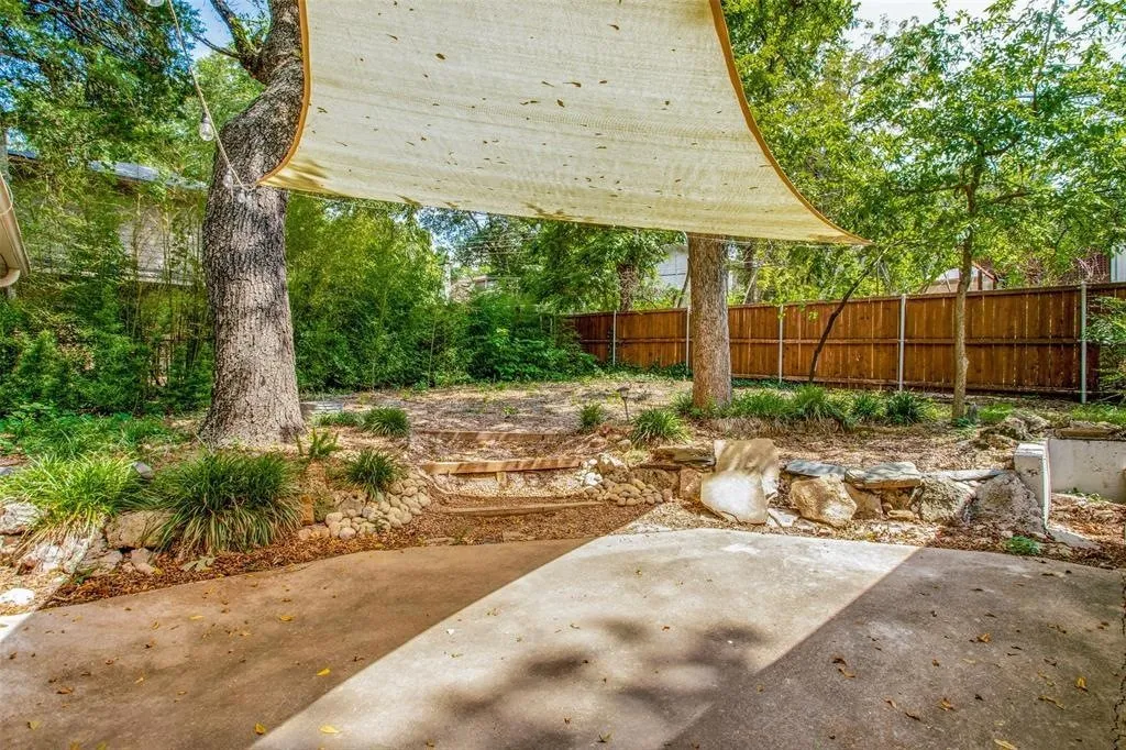 Concrete patio with an overhead shade sail, tiered landscaping, stone retaining walls, and a wood privacy fence