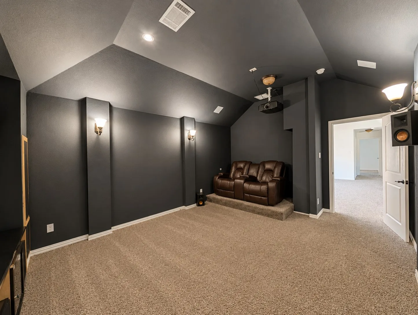 Carpeted entertainment space featuring sconce lighting, an elevated platform, and an integrated ceiling projector