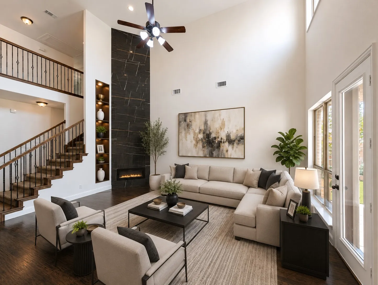Soaring ceilings, a two-story fireplace with integrated shelving and linear flame, wood-finish flooring, a two-story staircase with wrought iron balusters, and a ceiling fan with integrated lighting