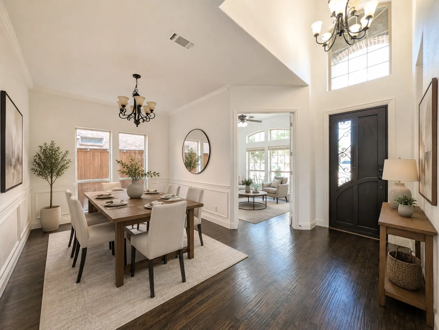 High ceilings, dark wood-finish flooring, two chandeliers, white wall paneling, and an arched entry door with decorative glass