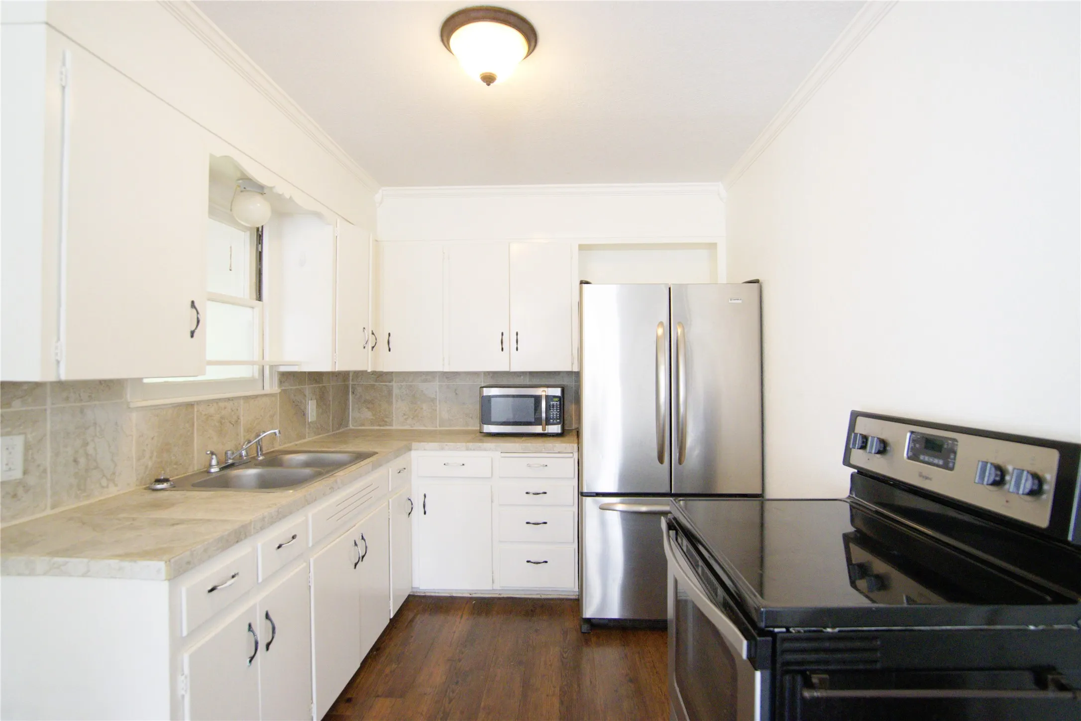 Galley kitchen featuring white cabinetry, stainless steel appliances including French-door refrigerator and electric range, tile counter and backsplash, viynl wood-finish flooring, and ample counter space with undermount sink