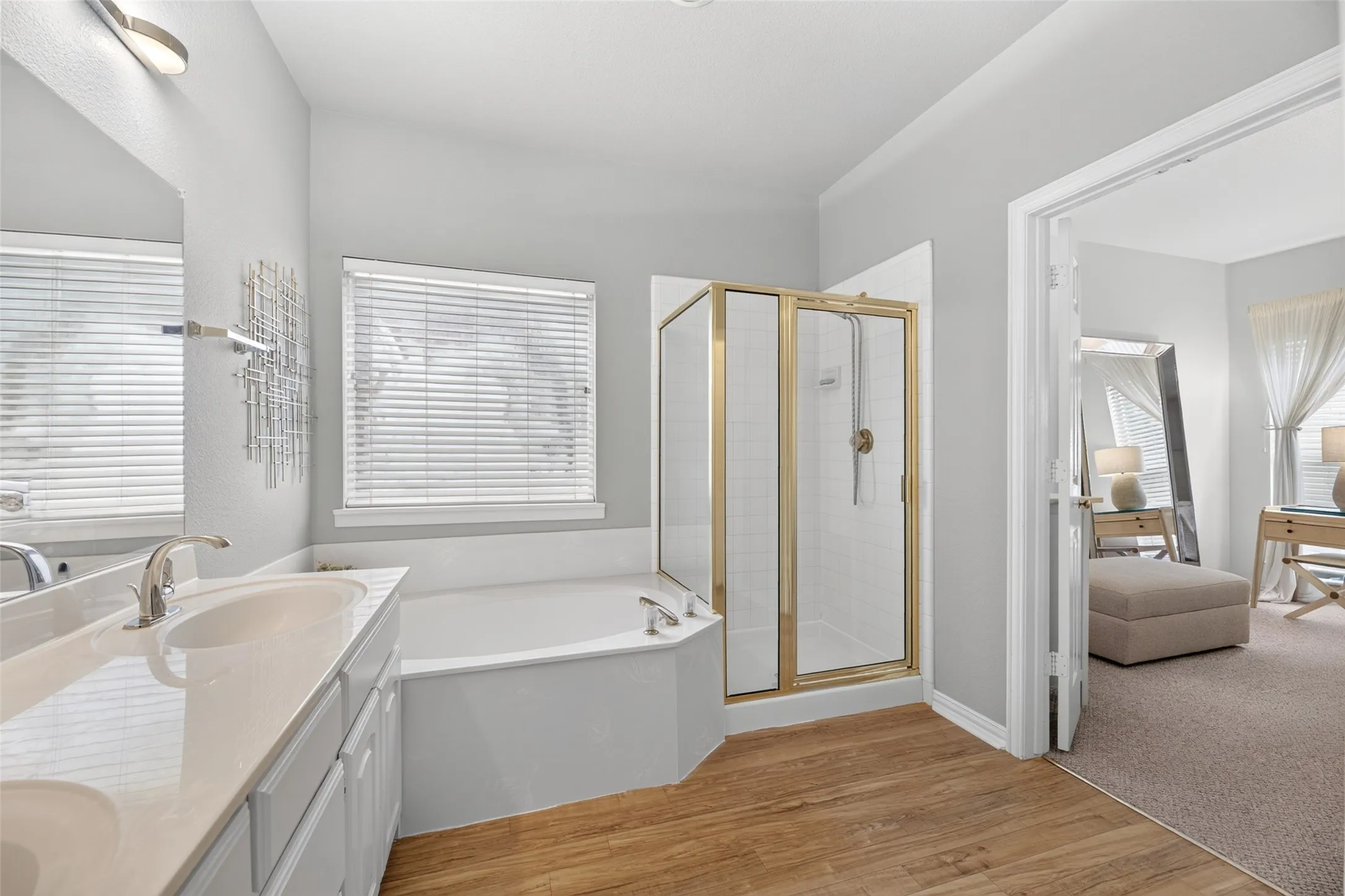 Bathroom featuring dual vanity with undermount sinks, wood-finish flooring, a corner soaking tub, and a glass-enclosed shower with gold-tone accents