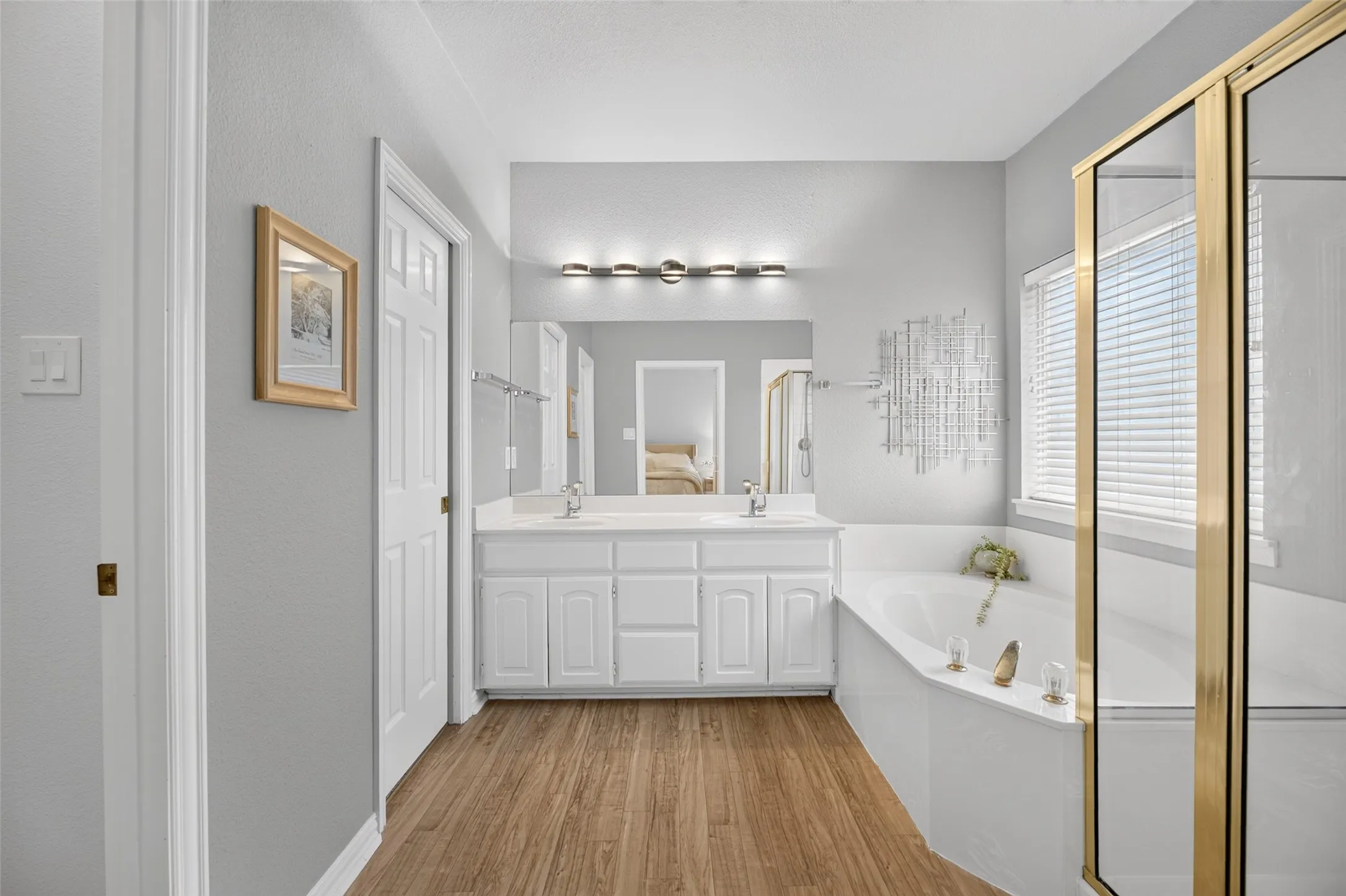 Dual-sink vanity with white cabinetry, wood-finish flooring, built-in bathtub, and an enclosed shower with gold-tone framing