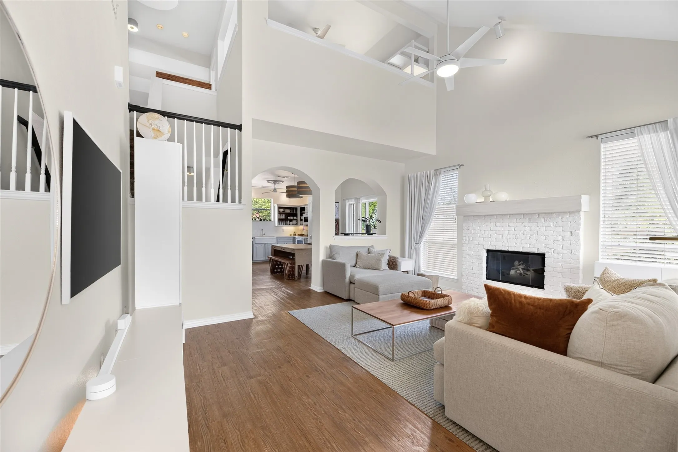 Vaulted ceilings, wood-finish flooring, a white brick fireplace, multiple windows with blinds, and arched doorways