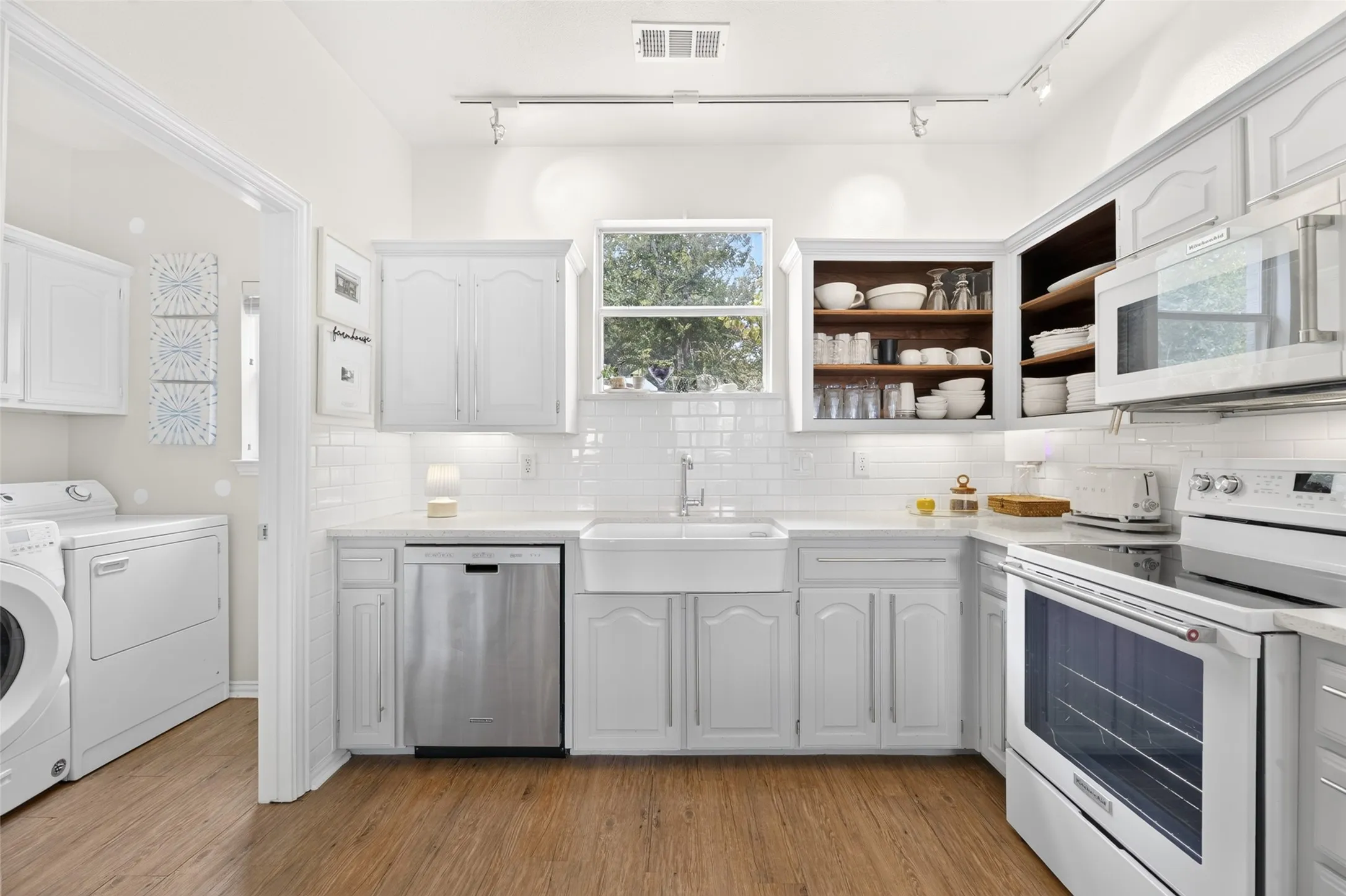 White cabinetry, subway tile backsplash, farmhouse-style sink, stainless steel dishwasher, and wood-finish flooring