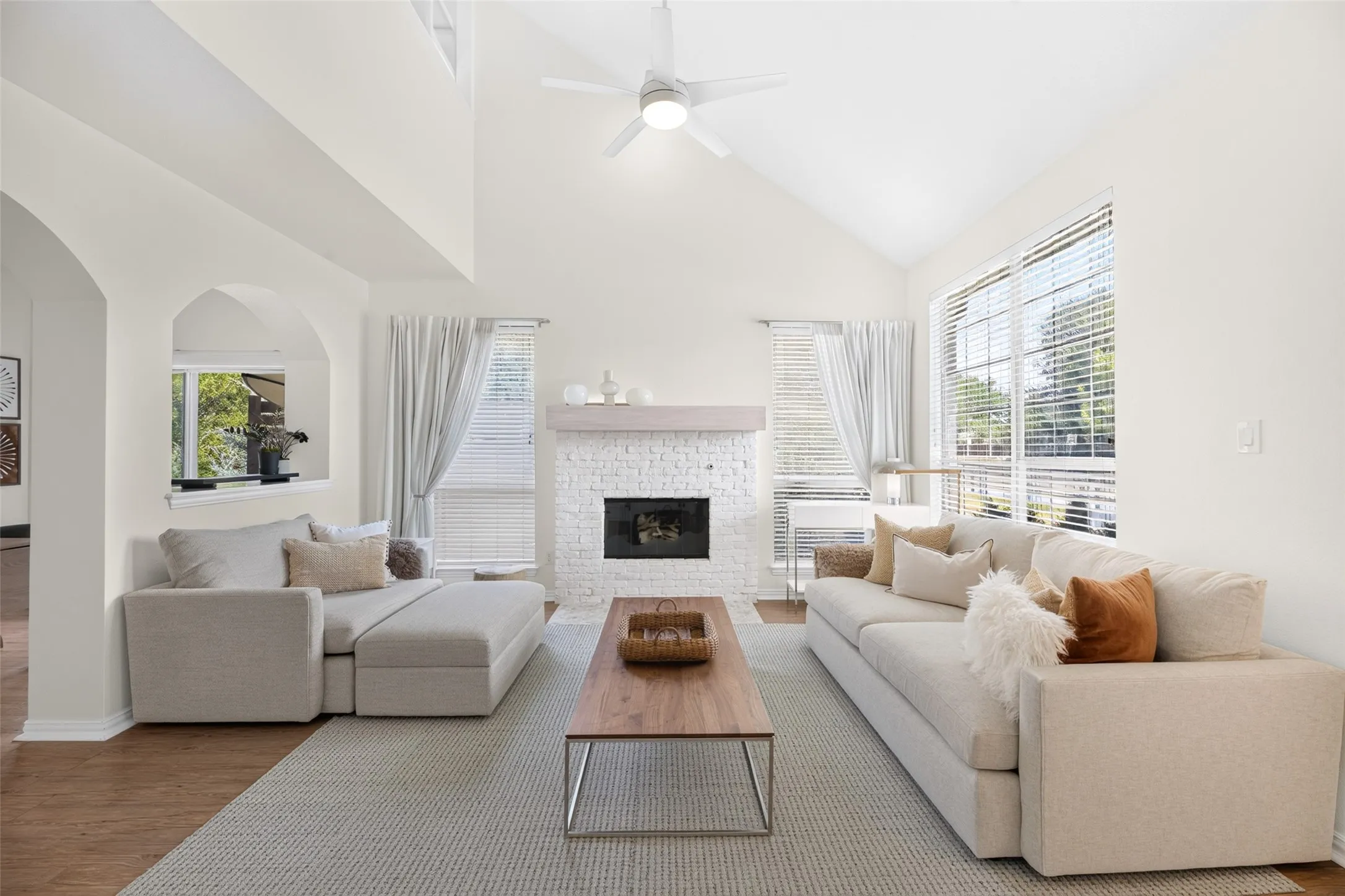 Vaulted ceiling, ceiling fan with light, wood-finish flooring, brick fireplace with mantel, and large windows