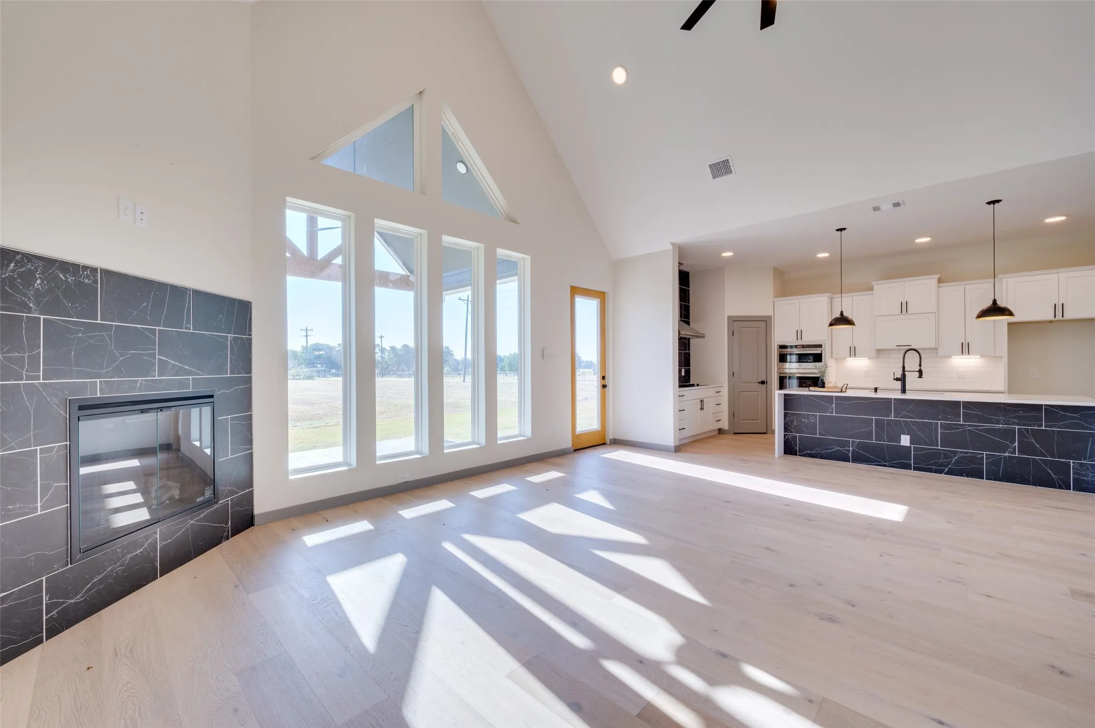 Vaulted ceiling with a ceiling fan, modern fireplace with a dark tiled surround, large windows, wood-finish flooring, and a kitchen island with dark tiled accents
