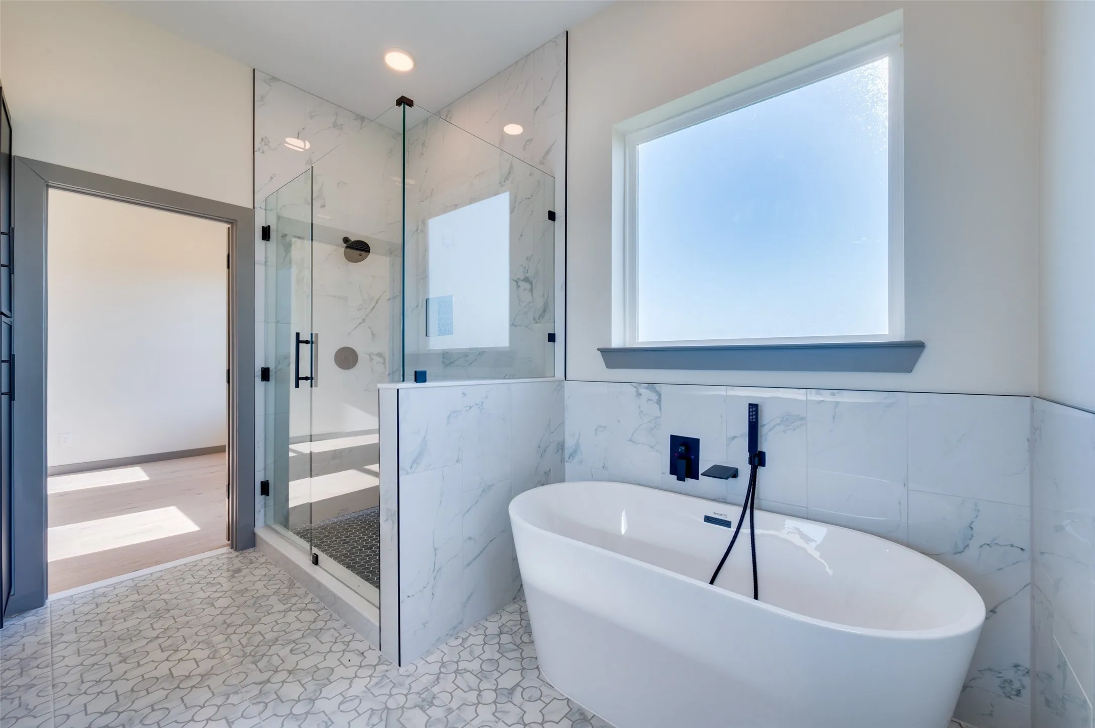 Freestanding soaking tub with matte black fixtures, glass-enclosed shower with stone-look tile walls, and patterned tile flooring