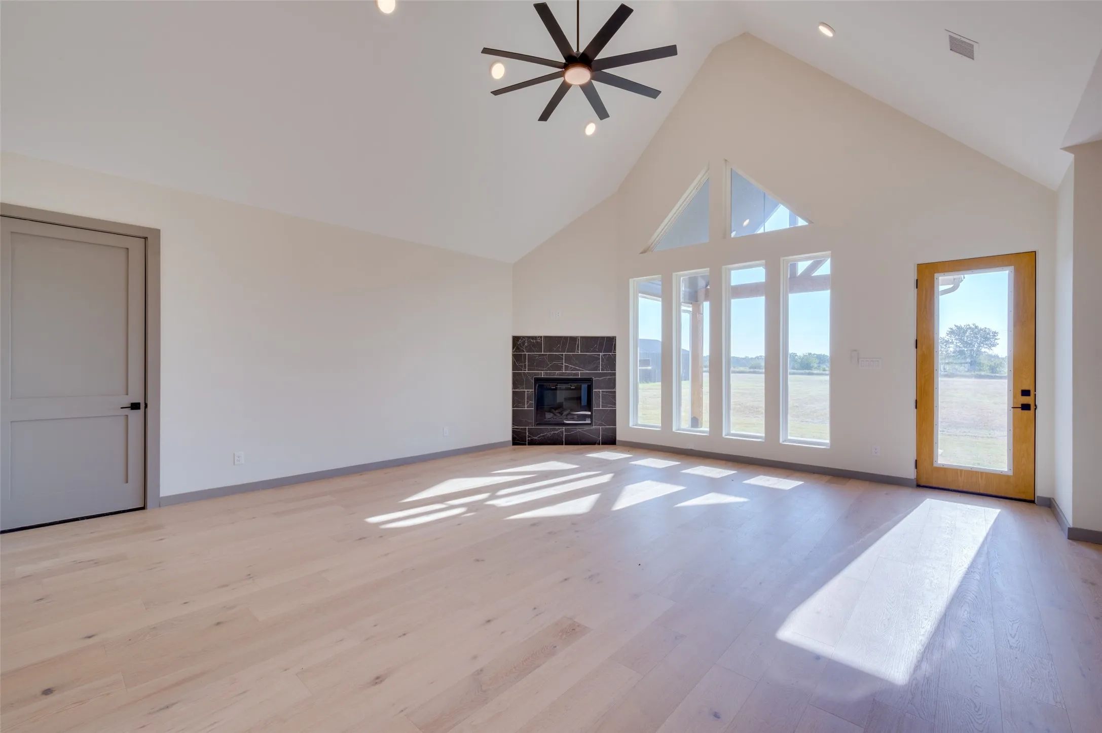 Vaulted ceiling with a contemporary ceiling fan, light wood-finish flooring, corner fireplace with dark stone surround, and expansive windows