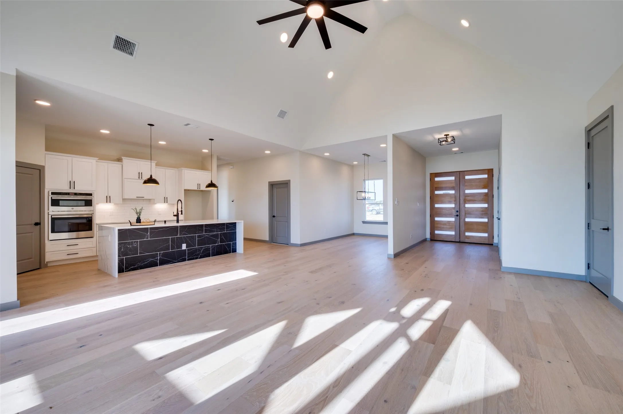 Vaulted ceilings with recessed lighting, light wood-finish flooring, a kitchen island with stone-finish paneling, white cabinetry, and a double-door entry