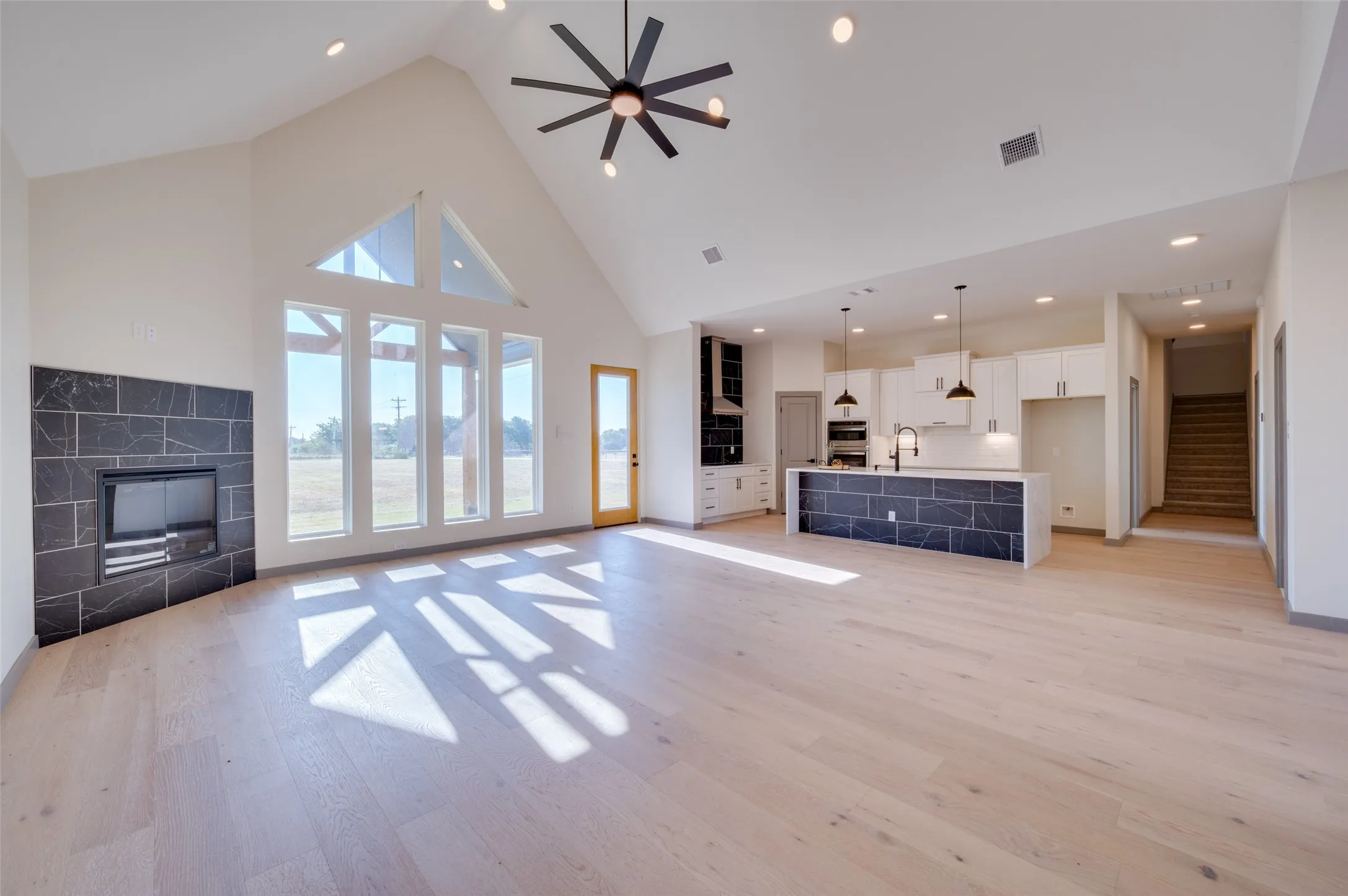Vaulted ceiling, wood-finish flooring, fireplace with dark tile surround, large windows, and a kitchen island with dark tile facade