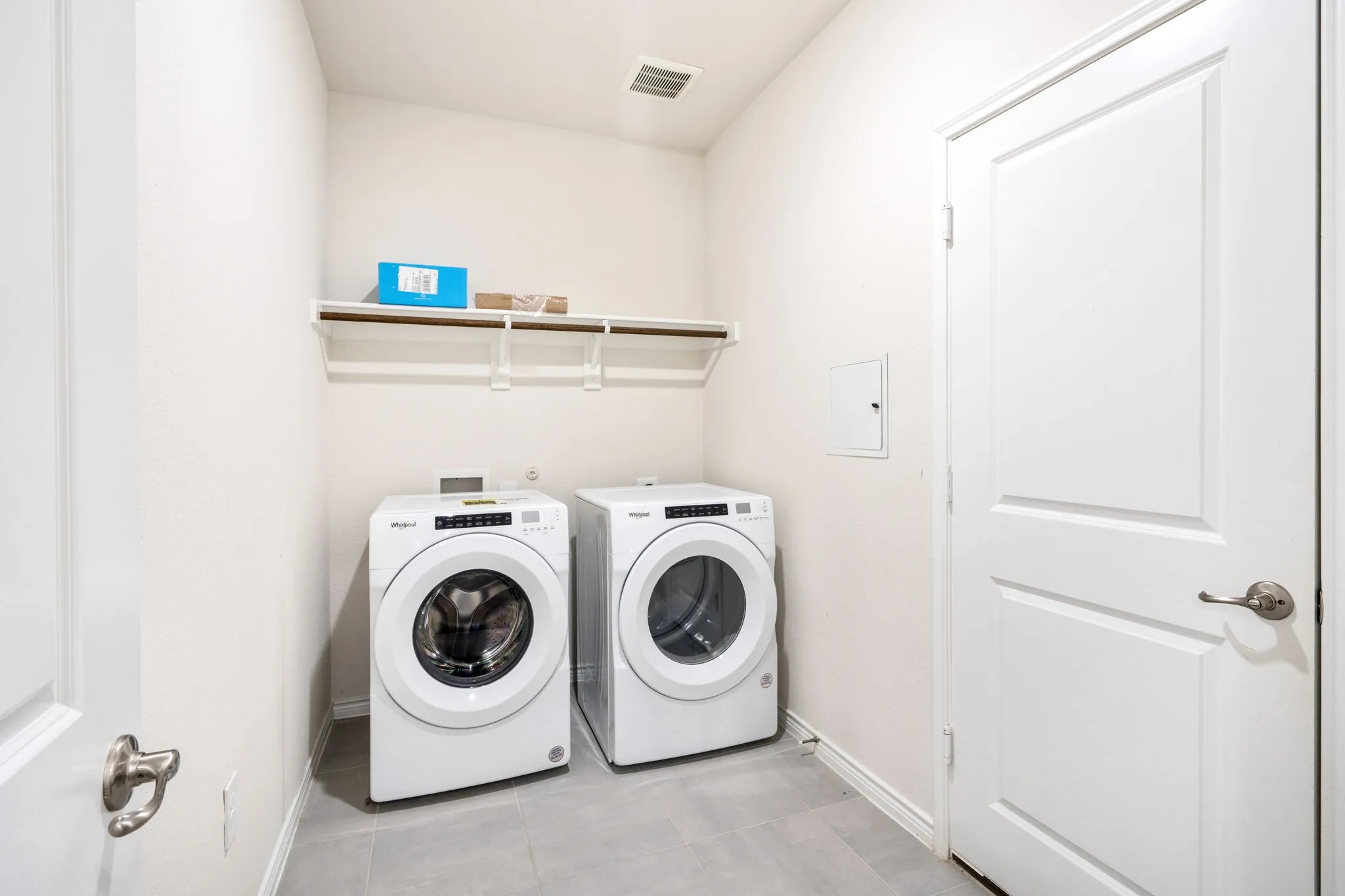 Laundry area with light gray tile flooring, white walls, and a mounted storage shelf