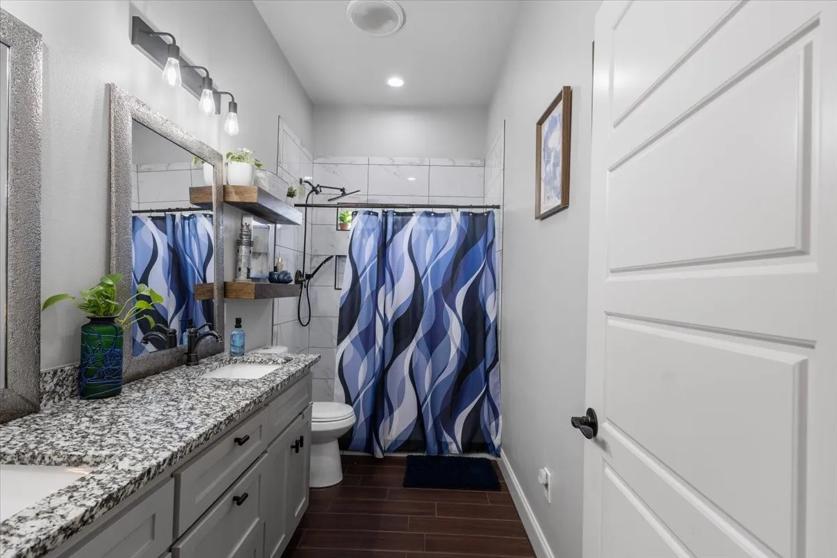 Dual vanity with granite countertop, wood-finish flooring, white shower tile, and a white interior door with contemporary paneling