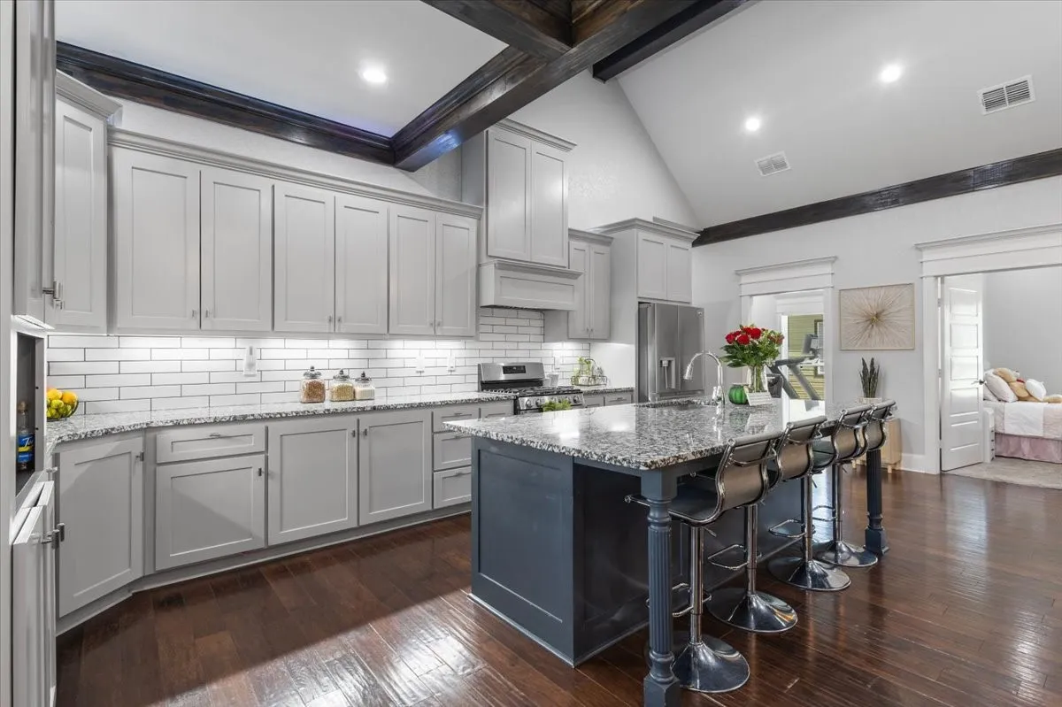 Kitchen featuring a vaulted ceiling with exposed beams, wood-finish flooring, a central island with a granite countertop, light-colored cabinetry, and a subway tile backsplash