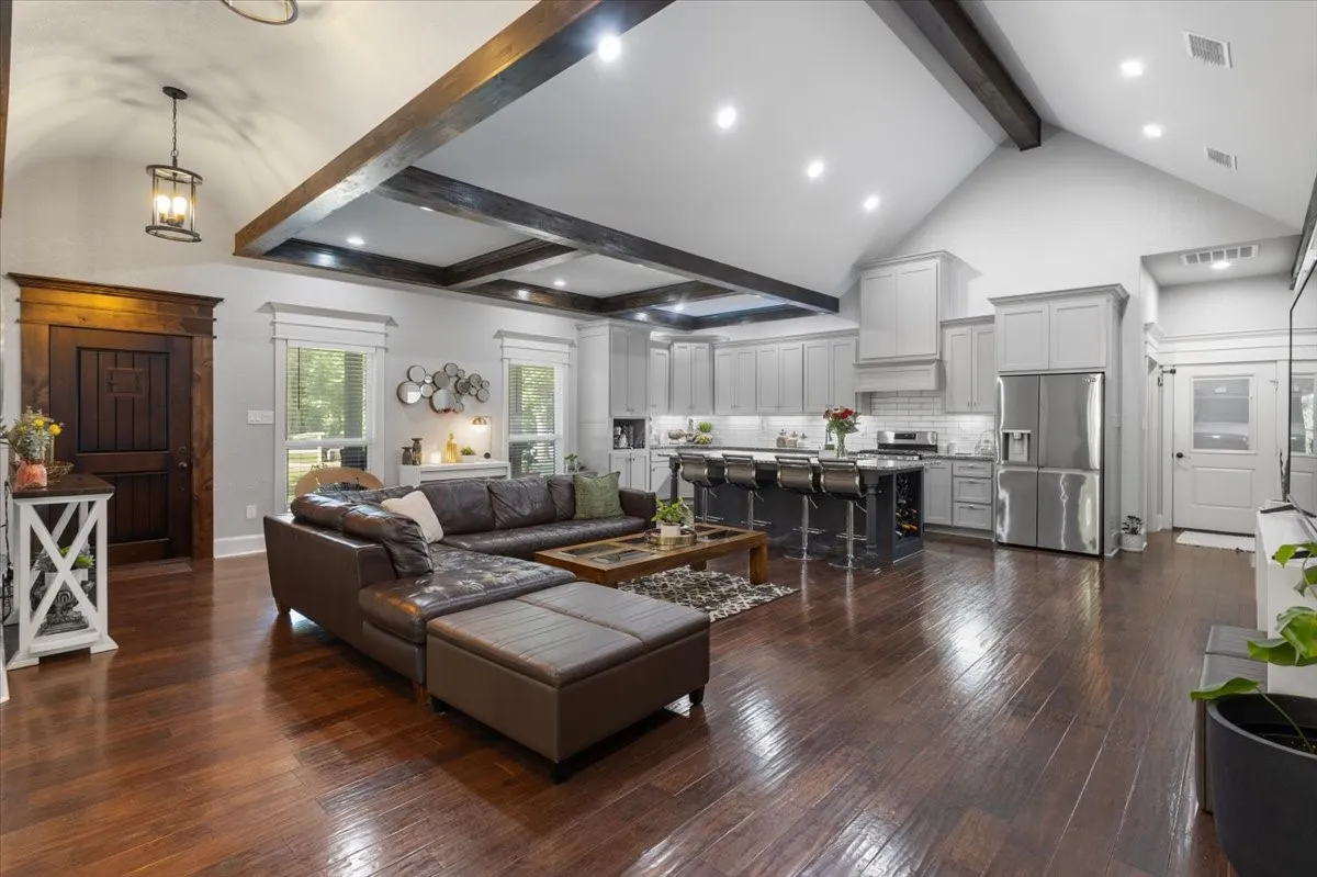 Vaulted ceiling with exposed wood beams, rich wood-finish flooring, a kitchen featuring stainless steel appliances, light gray cabinetry, and a dark island with seating