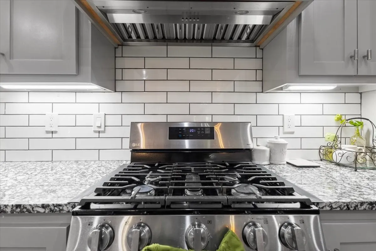 Stainless steel gas range with a coordinating range hood, white subway tile backsplash, light grey cabinetry, and speckled countertops