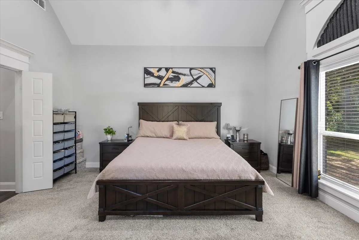 Vaulted ceiling, arched transom window, wood-finish bed frame, nightstands with drawer storage, and carpeted flooring