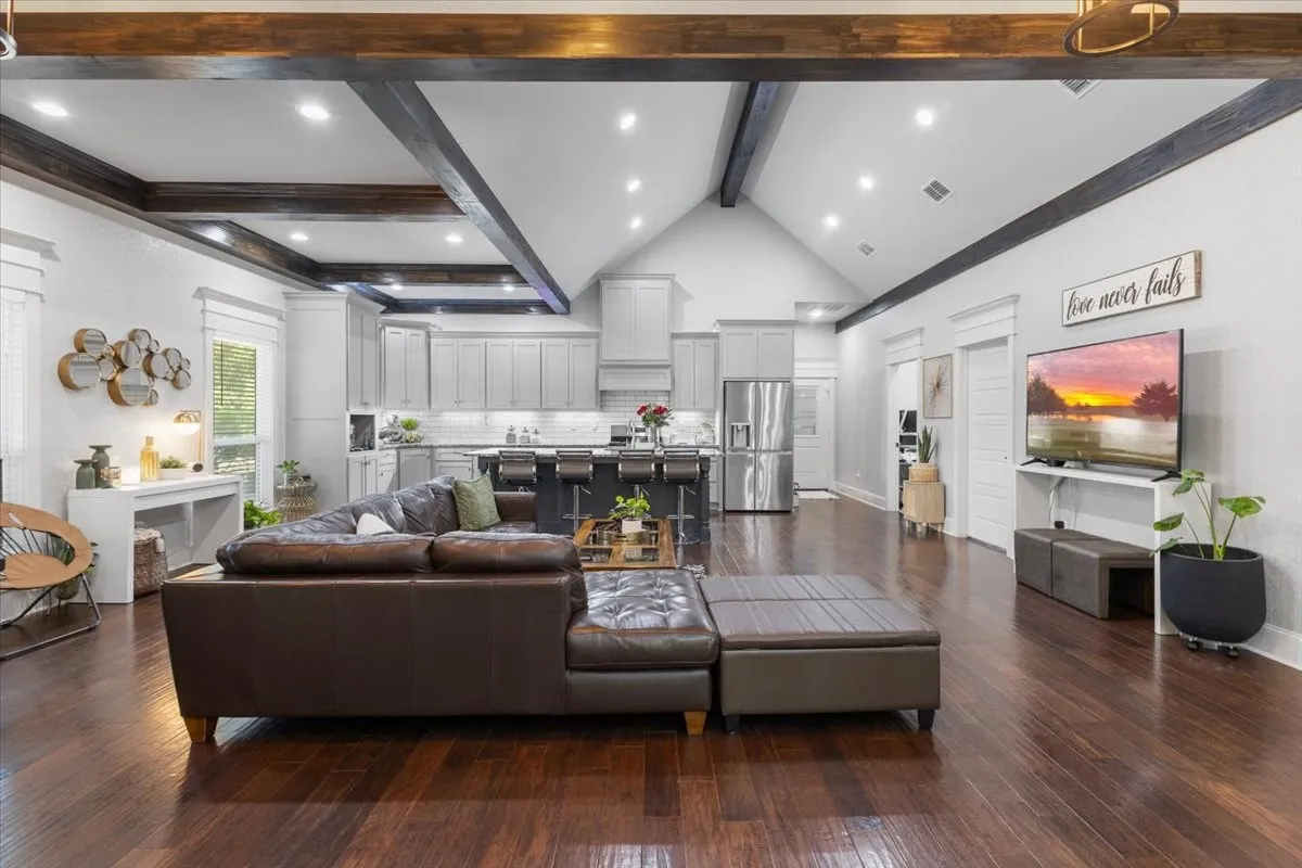 Open-concept living space with dark wood-finish flooring, vaulted ceiling with exposed beams, recessed lighting, and a modern kitchen featuring light gray cabinetry and stainless steel appliances