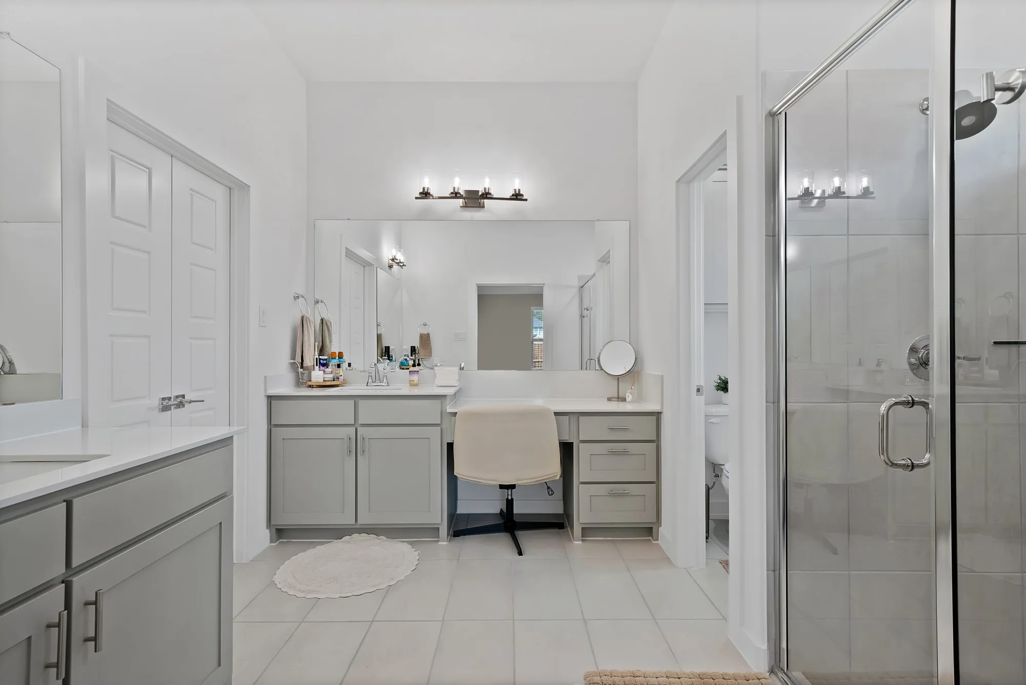 Dual vanity with white countertops and light gray cabinetry, glass-enclosed shower, tiled flooring, and a dedicated vanity seating area.