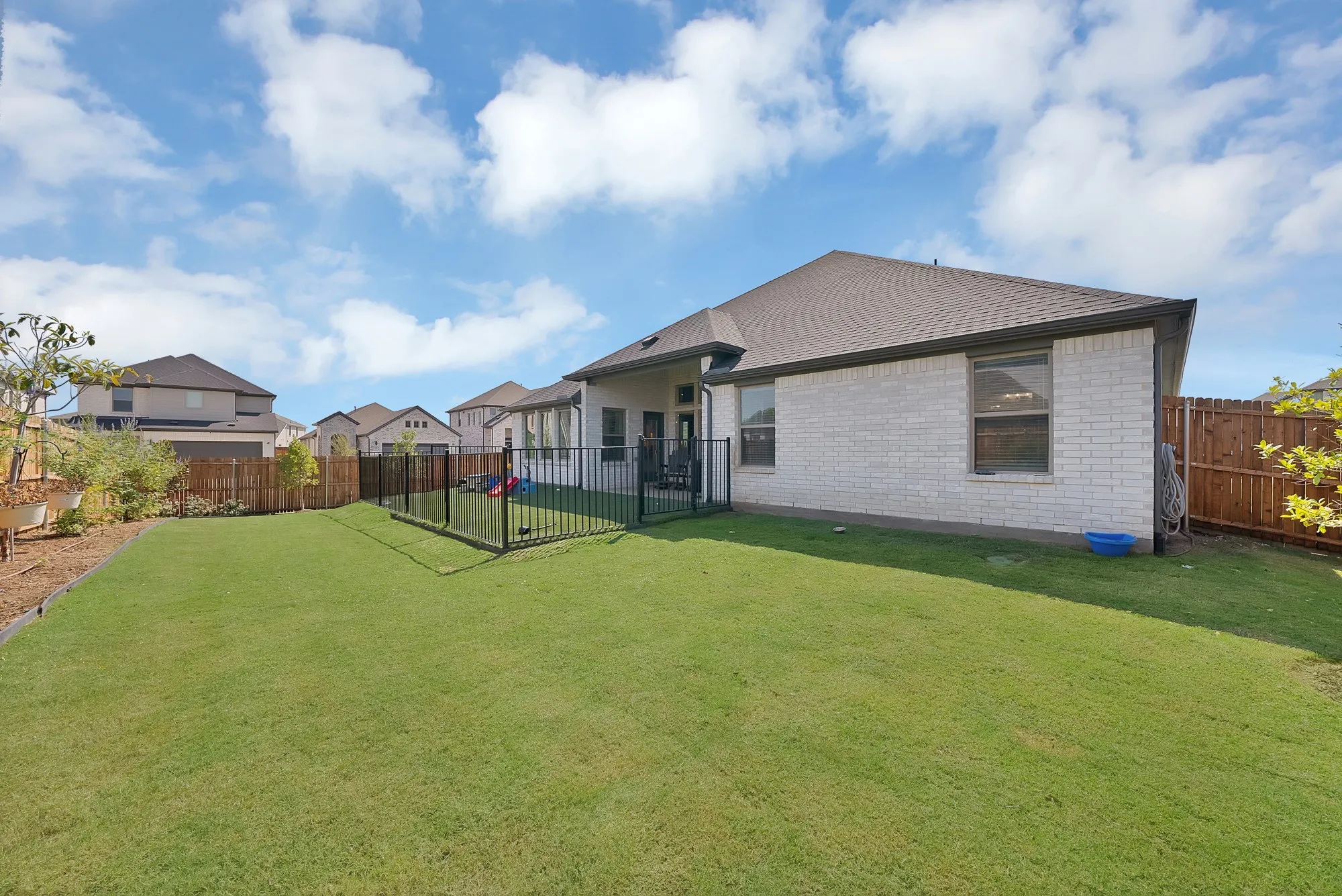 Expansive green lawn, white brick exterior, private wrought iron fenced area with artificial turf, brown wood perimeter fence, and professional landscaping that other homes do not have!