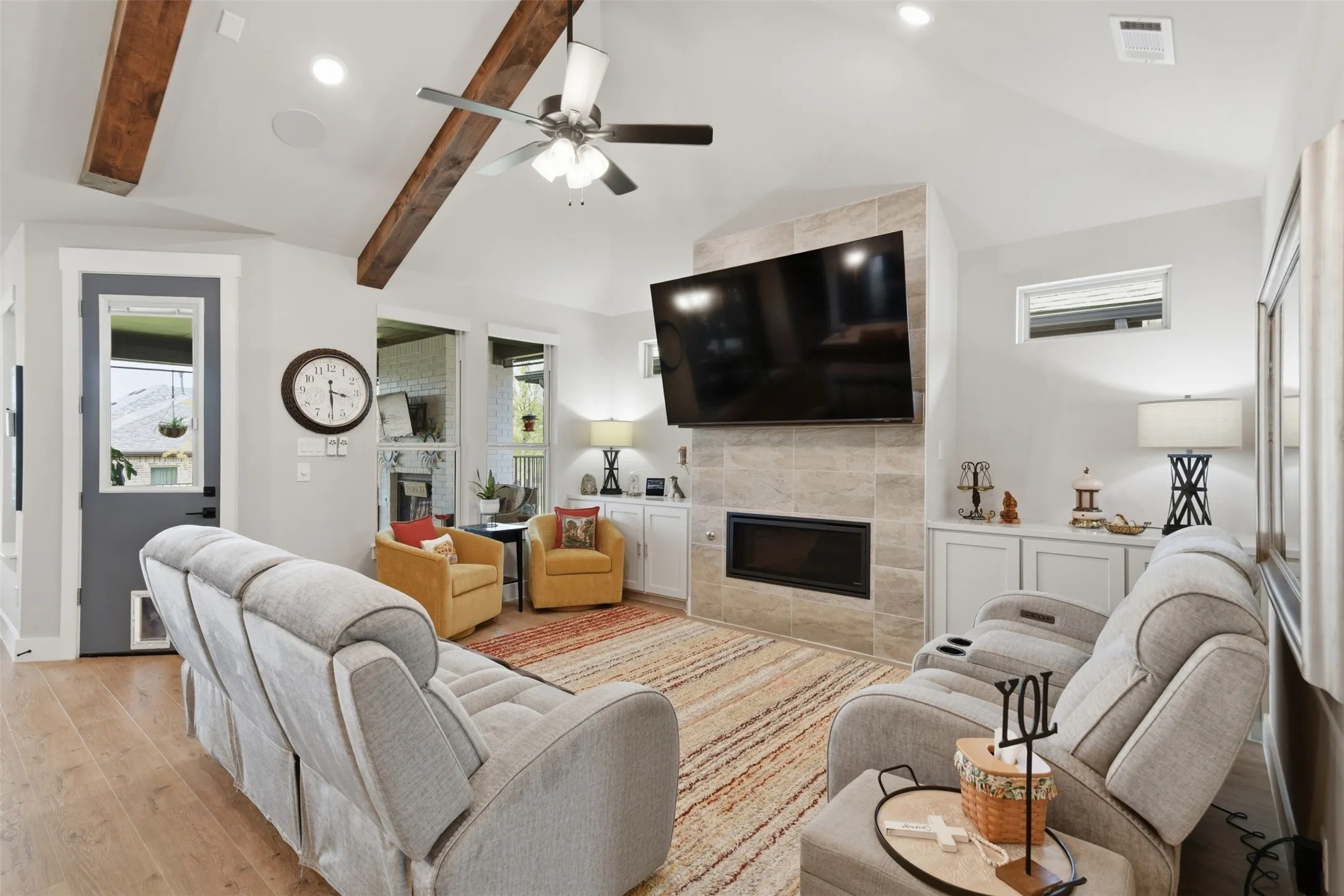 Vaulted ceiling with exposed wood beams, wood-finish flooring, built-in fireplace with tile surround, built-in cabinetry, and ceiling fan