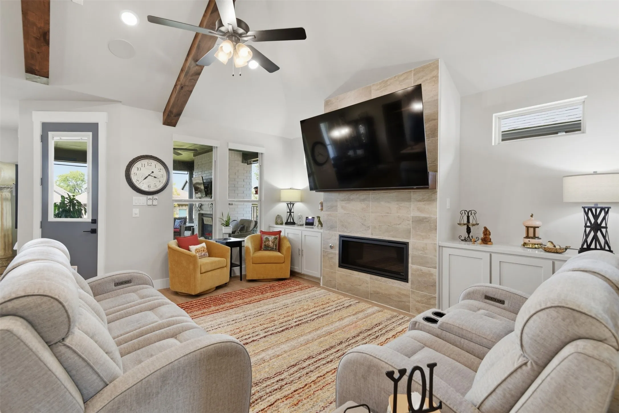 Vaulted ceiling with exposed wood beams, tile-clad fireplace, integrated built-in cabinetry, and ceiling fan with integrated lighting