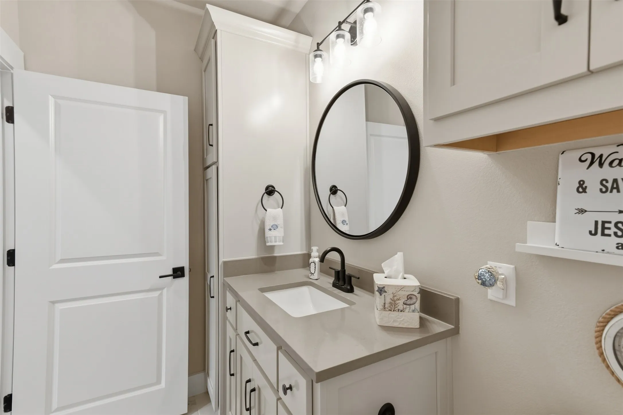 White shaker-style cabinetry with matte black hardware, integrated rectangular sink, stone-finish countertop, and a round black-framed mirror