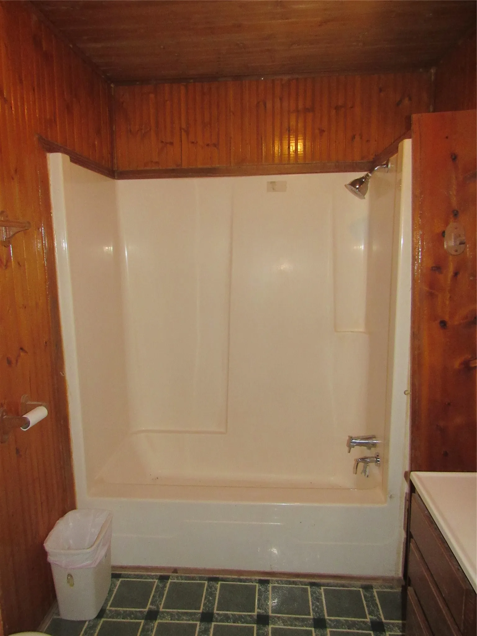 Wood-paneled walls and ceiling, full shower-tub combination with chrome fixtures, and a vanity with a light-colored countertop