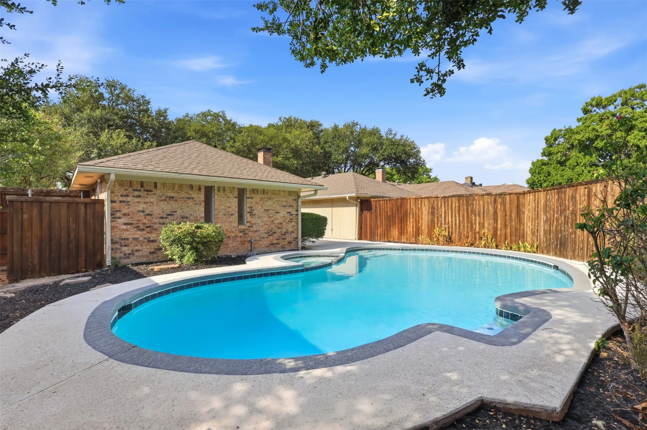 Freeform swimming pool with a concrete surround, wood privacy fencing, and brick exterior structures