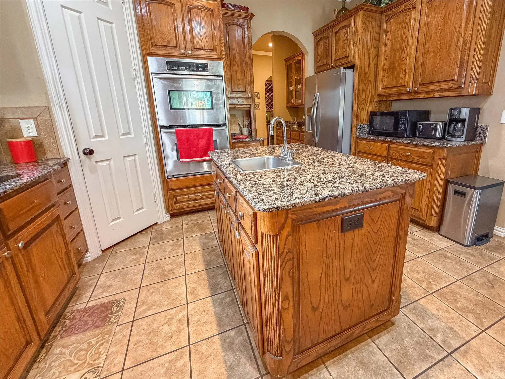 Kitchen featuring wood cabinetry, granite countertops, tile flooring, a center island with sink, and double wall ovens
