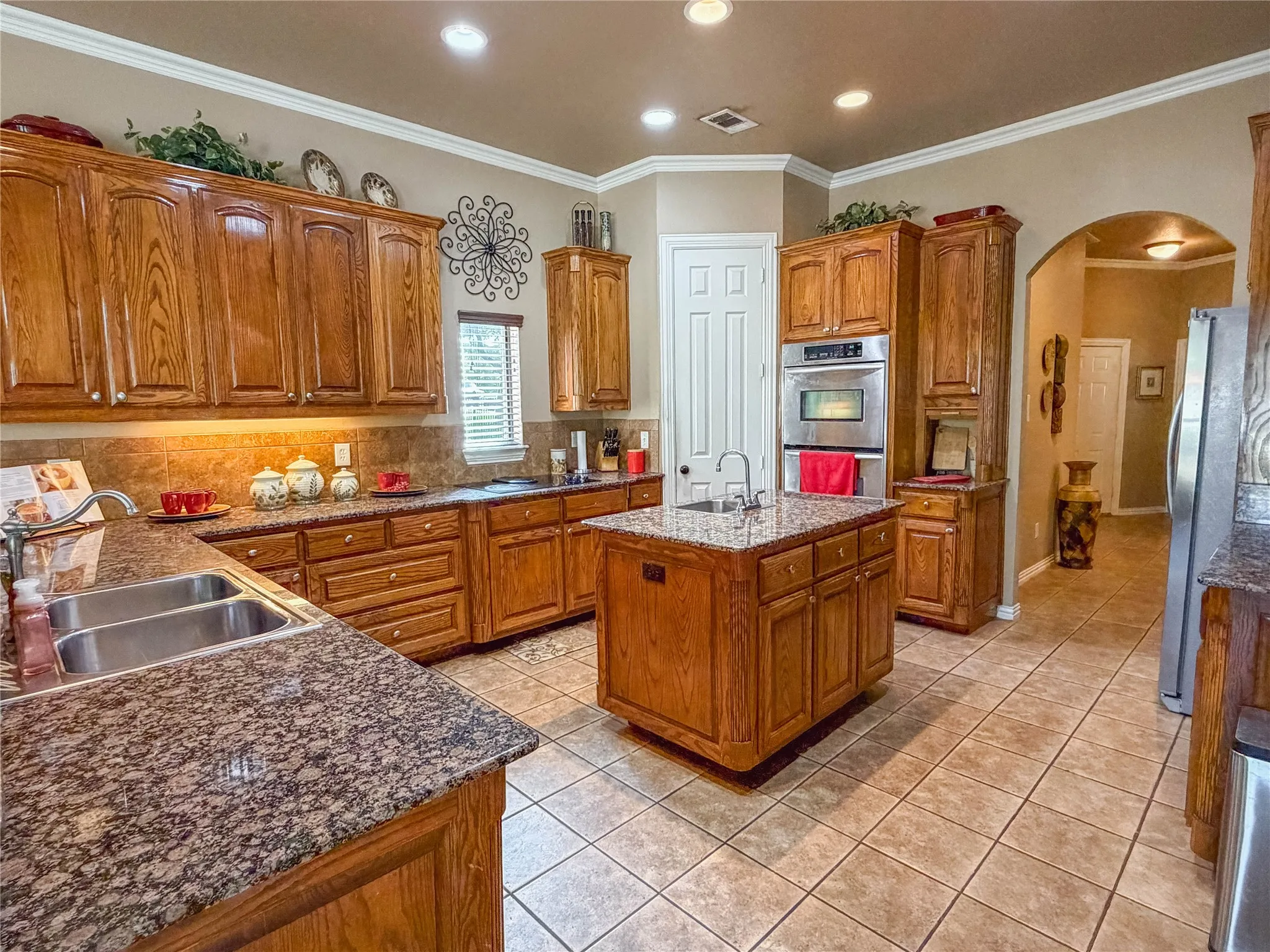 Spacious kitchen featuring rich wood cabinetry throughout, granite countertops, a large center island with seating, stainless steel appliances including a double wall oven, recessed lighting, and neutral tile flooring